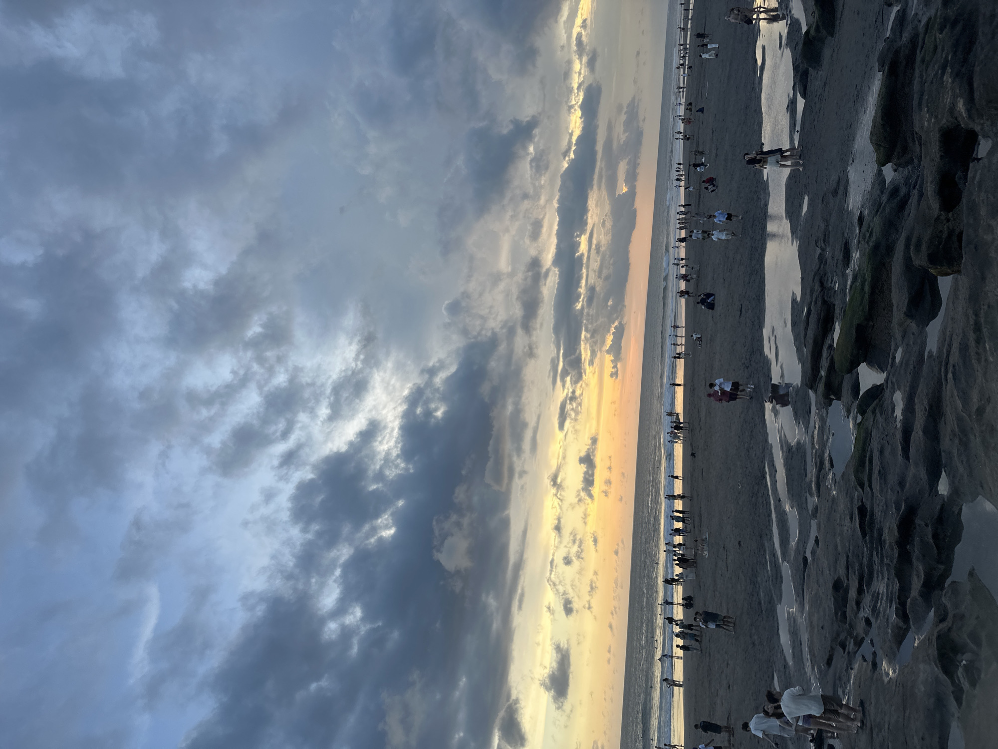 The image depicts a beach scene at sunset in Canggu, Indonesia. A crowded shoreline stretches along the horizon, with numerous people visible on the sand. The foreground contains dark, rocky formations, some reflecting the sunset's light. The sky is dominated by heavy, overcast clouds, transitioning from blue-gray tones to a vibrant yellow-orange near the horizon where the sun is setting. The orientation of the image is rotated 90 degrees counter-clockwise.