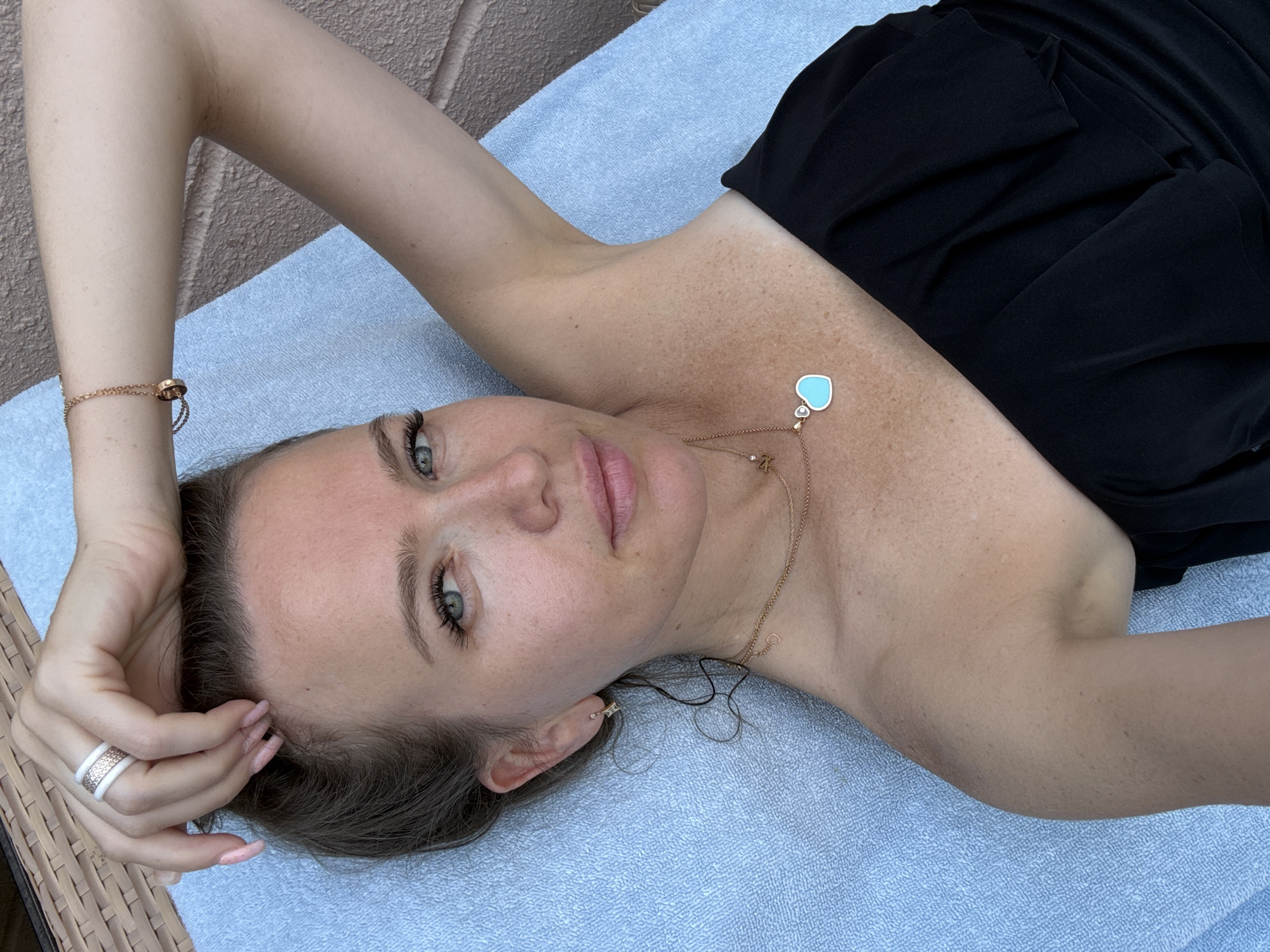A woman with blonde hair and blue eyes is lying on a light blue towel. She is wearing a black strapless top. She has freckles on her arms and chest. She is wearing jewelry, including a ring on her left hand, a bracelet on her right arm, and a necklace with a heart-shaped pendant. Her right arm is raised and her hand is resting on her head. The background is blurry and appears to be an outdoor setting with a textured surface that could be a wall or a lounge chair. The lighting suggests it is daytime, possibly sunny.
