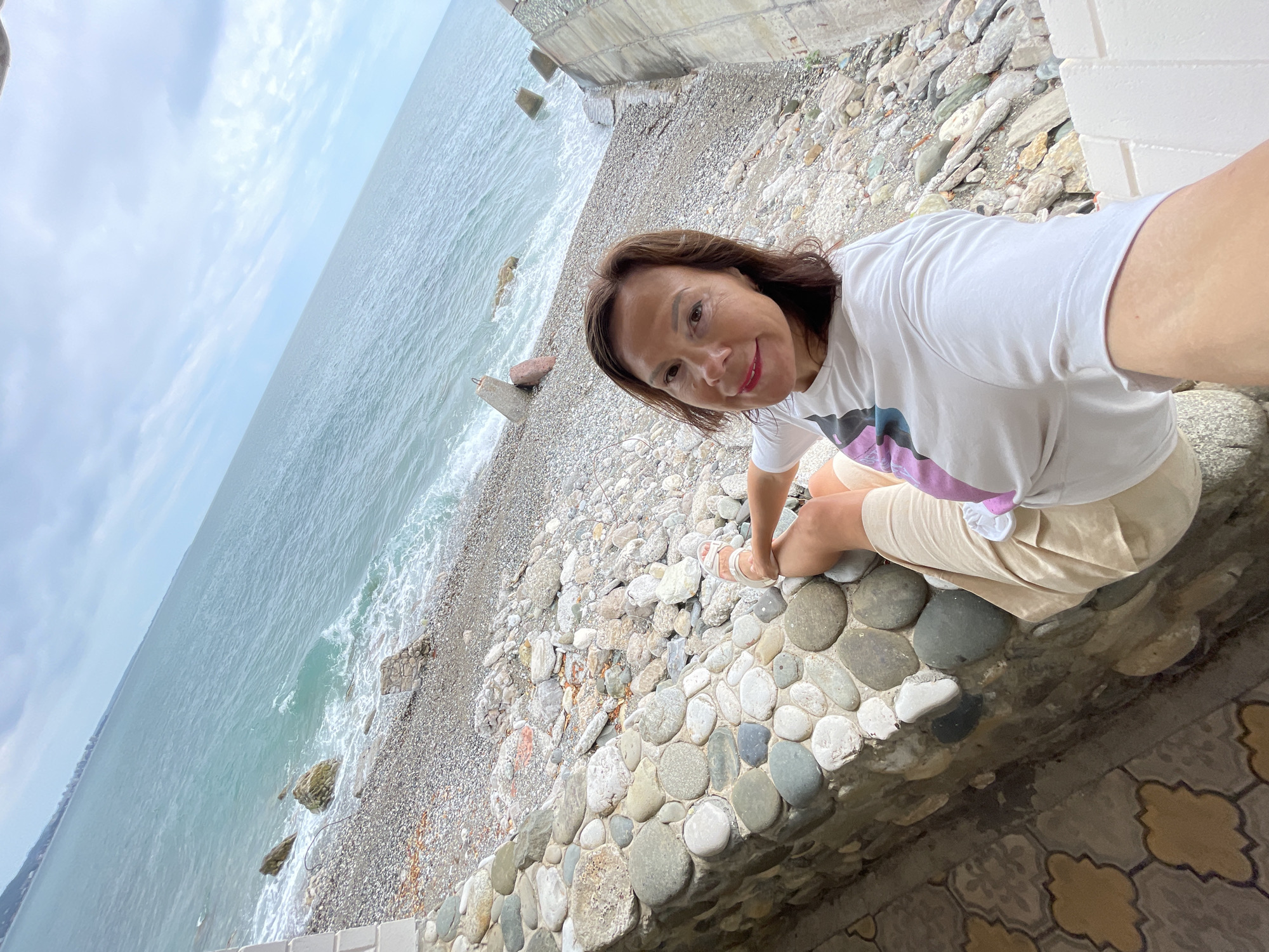 A woman with shoulder-length brown hair and dark eyes is taking a selfie on a pebbly beach in Abaata, Abkhazia. She is wearing a white t-shirt with a colorful abstract design on the front and light-colored shorts. Her left leg is bent with her foot resting on a low stone wall made of rounded grey and white stones. The beach is covered in small grey and white pebbles, with some larger rocks and concrete blocks scattered along the shoreline. The turquoise water of the sea is visible in the background, with waves breaking on the shore under a cloudy sky. To the left of the woman, a patterned tiled surface is visible.