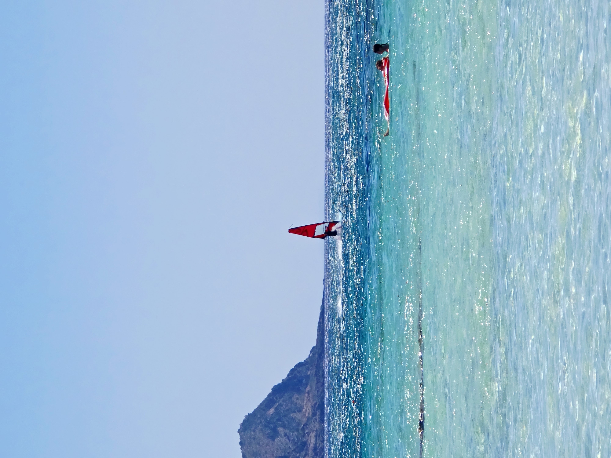 This is a photograph taken on a sunny day in Greece, showing a vibrant blue sky meeting a sparkling turquoise sea. The water is clear and shallow near the shore, gradually deepening into a richer blue. In the distance, a lone windsurfer with a red sail navigates the waves. To the right, two individuals are enjoying the water; one is lying on an orange inflatable raft, while the other appears to be swimming nearby. A rocky island or promontory is visible on the horizon, adding a dramatic backdrop to the scene. The overall impression is one of a peaceful and idyllic beach day, with a sense of both activity and relaxation.