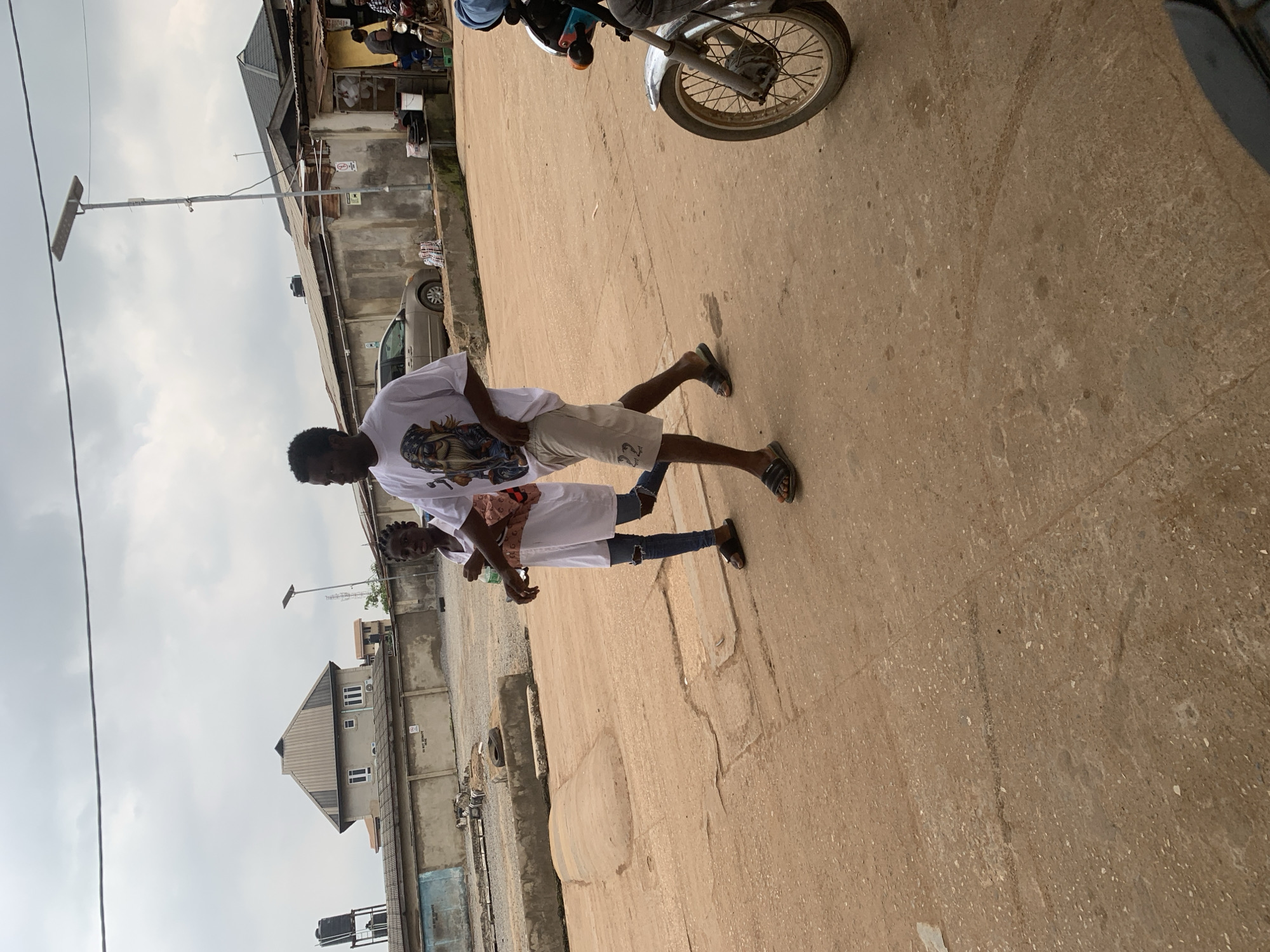 The image captures a scene on a street in Shagamu, Nigeria. Two individuals, a man and a woman, are walking side-by-side on a dusty, paved surface. The man, on the right, is wearing a white t-shirt with a graphic print and light-colored shorts. He has dark hair and is looking forward. The woman, to his left, is wearing a white t-shirt dress and ripped blue jeans. She has dark hair styled in braids and is also looking forward. A motorcycle is partially visible on the right side of the frame, with its front wheel and a rider wearing a blue helmet. In the background, there are buildings, some with corrugated metal roofs, and a solar-powered street light. The sky is overcast. The environment appears to be an urban or semi-urban area.