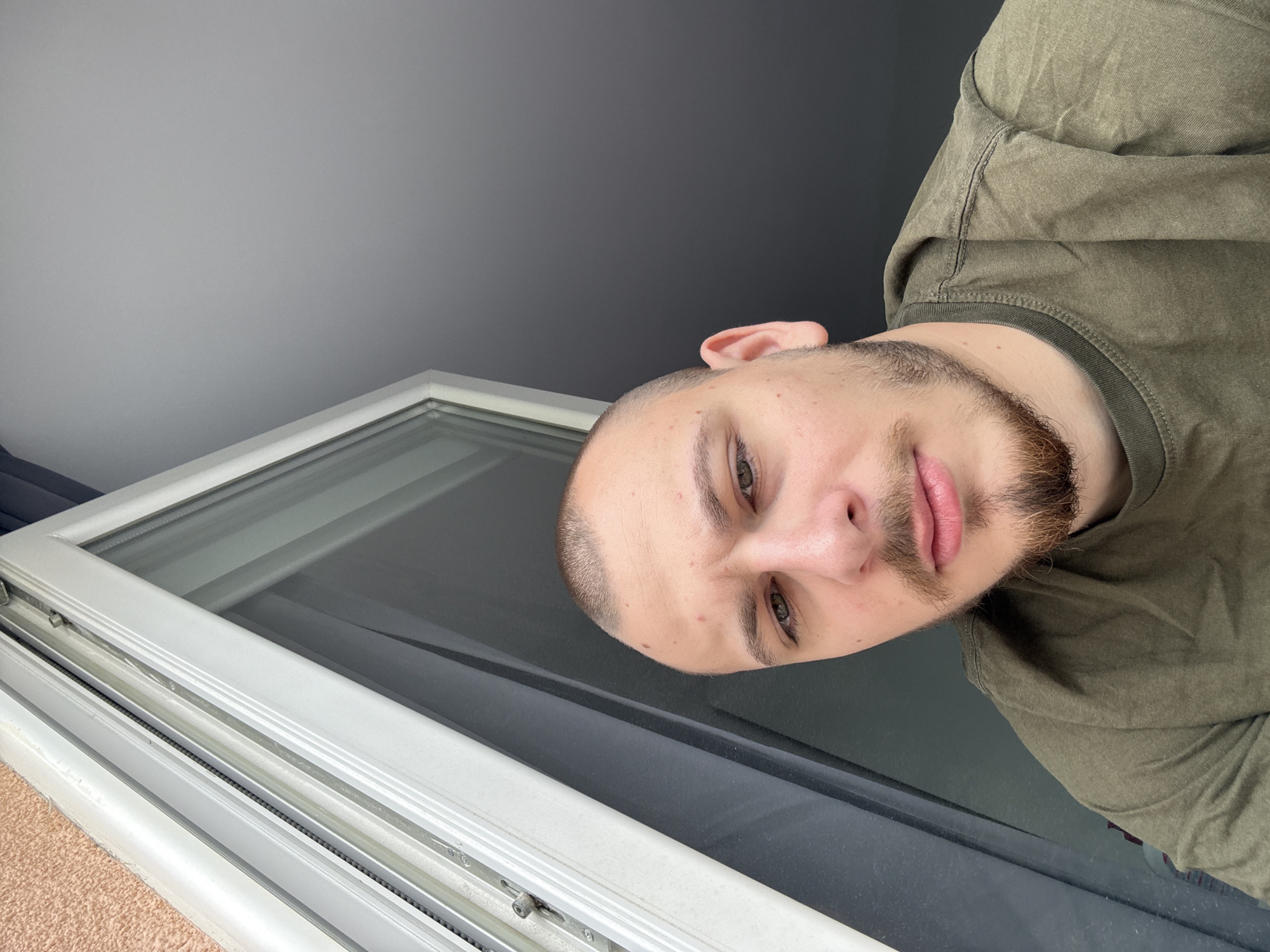 A man with a close-cropped haircut and a beard is looking down at the camera. He's wearing a green t-shirt and standing next to a white-framed window. The wall behind him is gray. The location cannot be determined from the image provided.