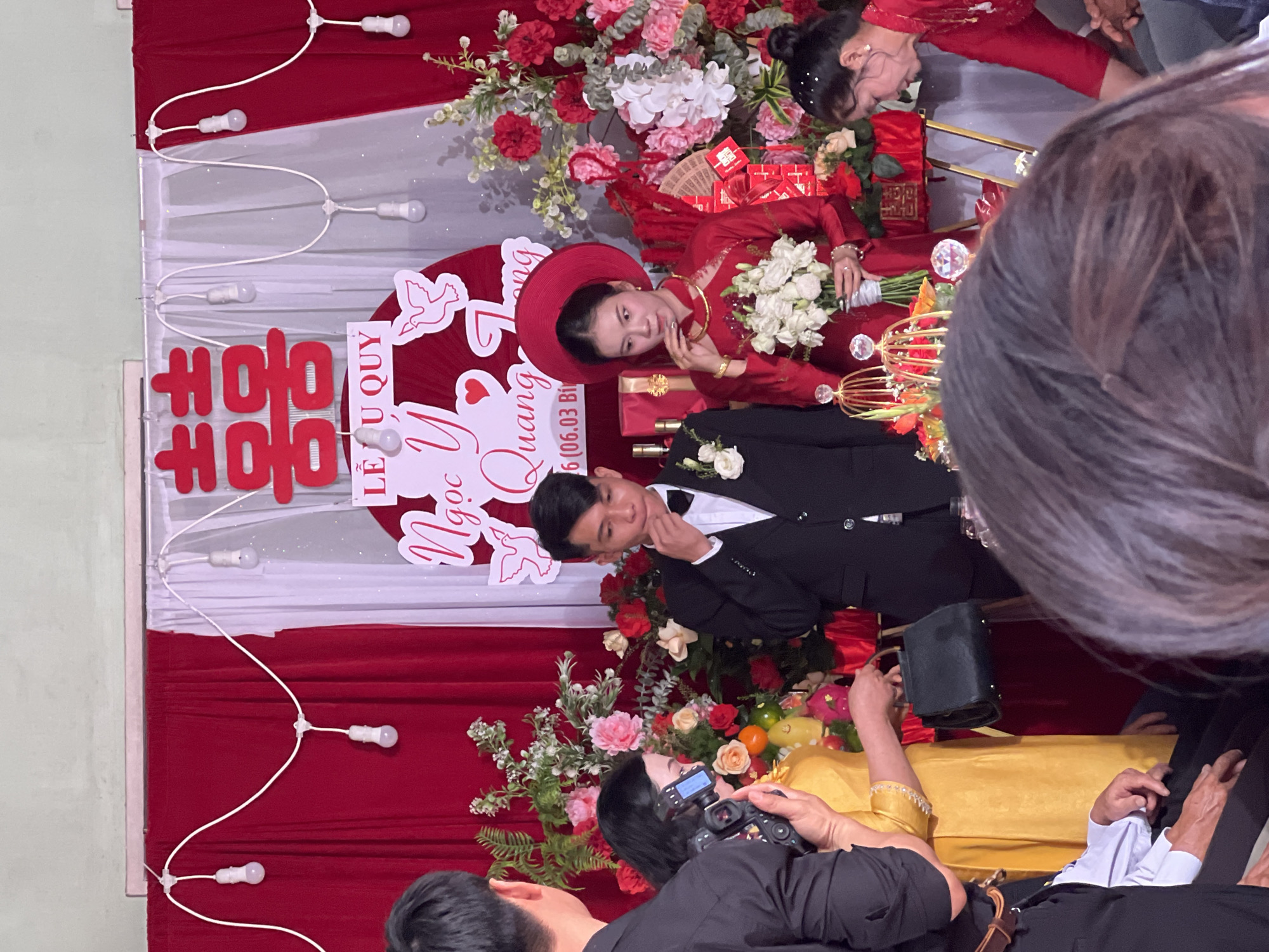 The image shows a wedding scene, possibly in Tay Son Commune, Vietnam. A groom in a black suit and white shirt looks at the bride, who is wearing a red dress and a traditional red hat. She is holding a white bouquet. Behind them is a red and white backdrop with decorations and the names "Ngọc Y" and "Quang" along with "LỄ U QUY" and the date "16.06.03 BÌ". There are strings of lights along the backdrop. Several people are in the scene, including a photographer taking pictures of the couple. The environment includes floral arrangements with red, white, and pink flowers.