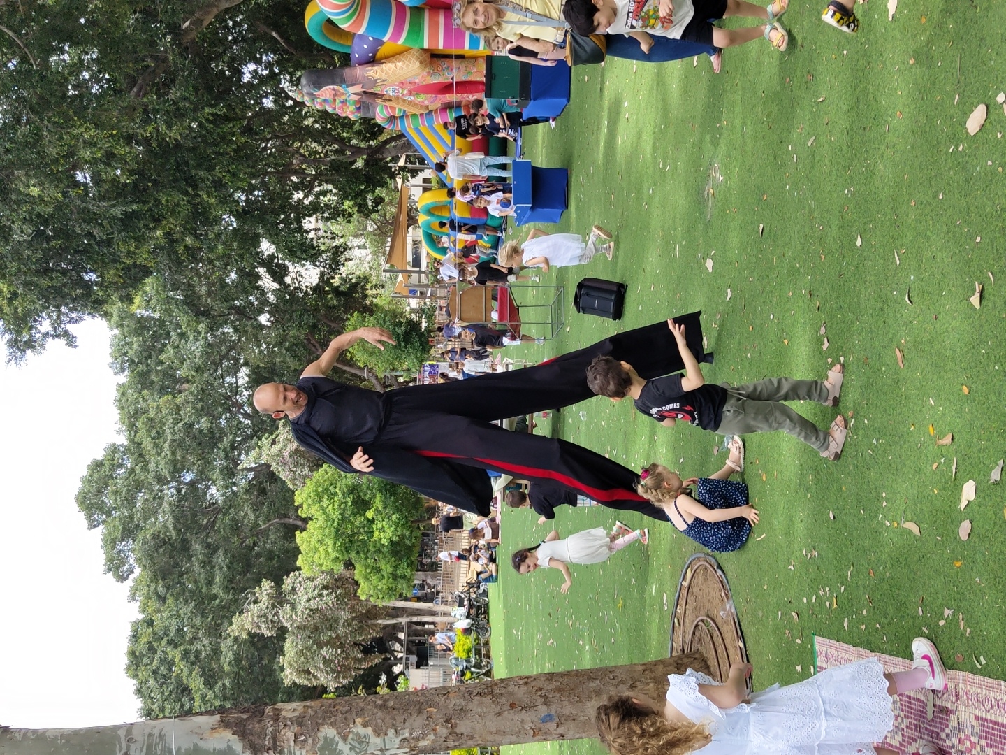 The image captures an outdoor event at a park in Tel Aviv, Israel, with a variety of elements suggesting an entertainment or festival atmosphere. The scene is set on a grassy area, partially shaded by large trees.
A performer on stilts is the central figure, dressed in a black outfit with red stripes down the sides of the legs. He appears to be interacting with a group of children. Some of the children are scattered around on the grass. A few are near the performer, looking up at him. Other children are standing and watching.
In the background, there are more people seated and standing, suggesting an audience. Further back, there are inflatable structures that appear to be part of the event's setup. Other identifiable objects include seating and what appears to be audio equipment.
The environment is typical of a park setting, with trees providing shade. The overall impression is one of a community event or performance in a public park.