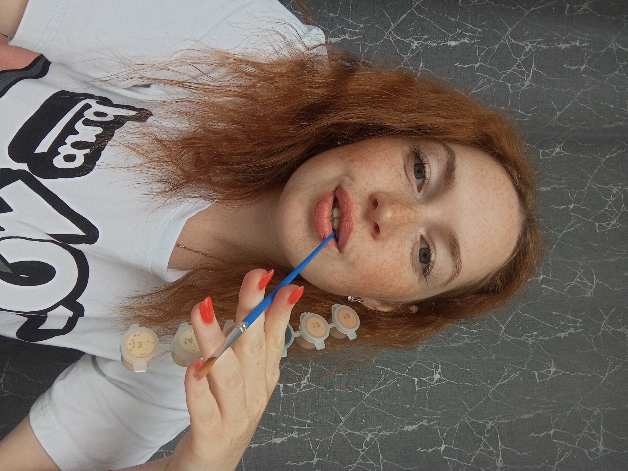 A person is seen from a high-angle view, seemingly lying on a surface with a grey, marble-patterned texture. The person has red hair and freckles on their face. They are wearing a white t-shirt with a black and white design. They are holding a blue paintbrush, with the bristles near their lips. Several small, plastic containers with numbered labels are present near their hand. The location, Yurgamysh, Russia, cannot be independently verified from the image.