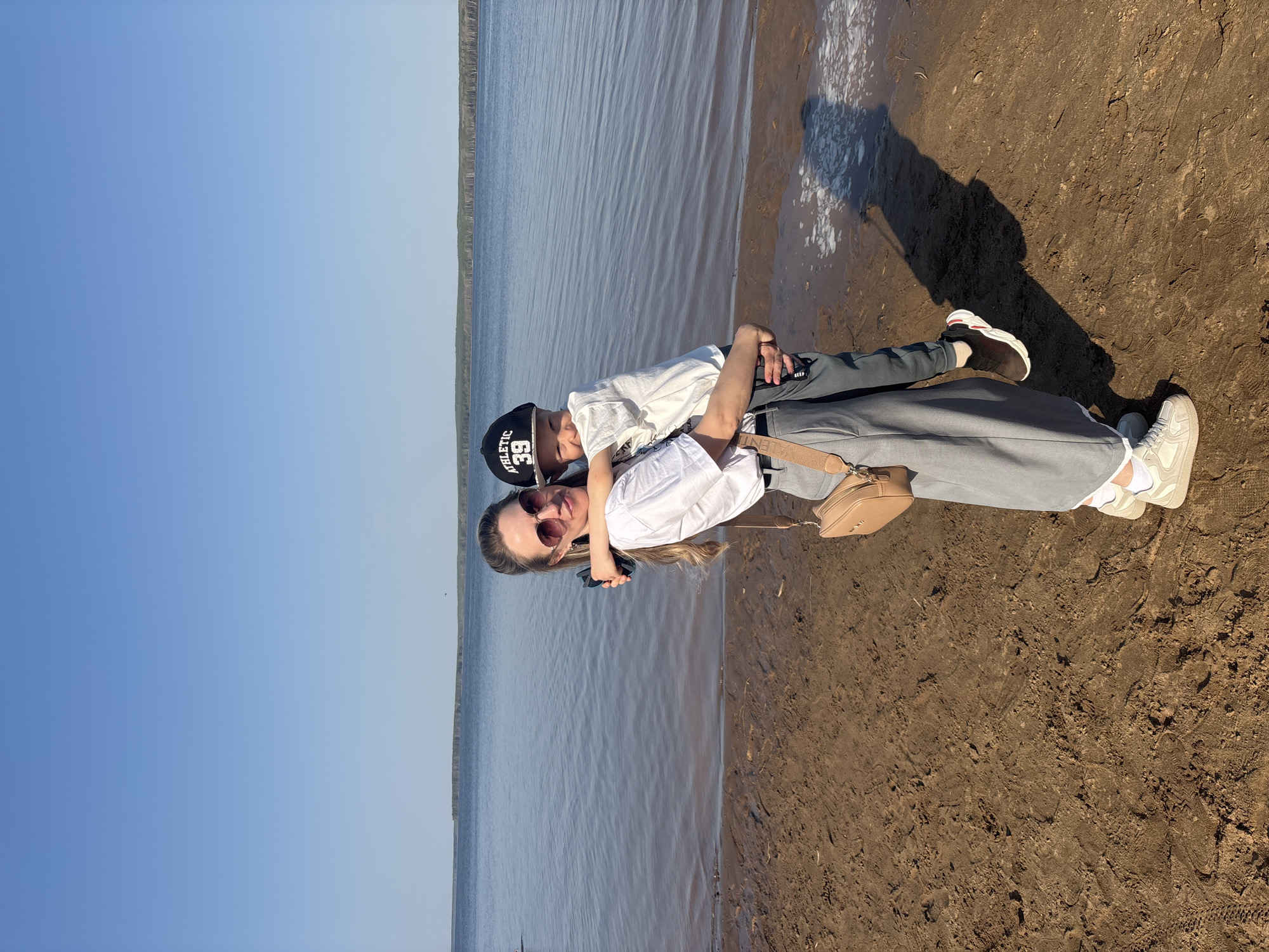 A photo shows a woman and a child on a beach in Chaikovsky, Russia. The woman is wearing sunglasses, a white shirt, and gray pants, and has a beige purse. The child wears a baseball cap, a white shirt, and gray pants. They are near the water's edge, with the lake extending to the horizon. The sky is blue. The beach is sandy and muddy, marked with footprints.