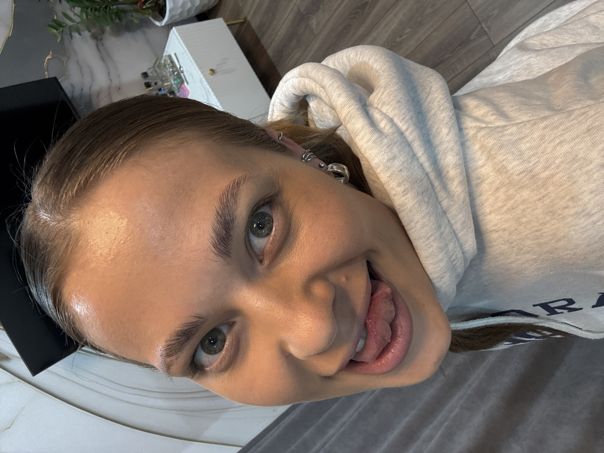 An indoor, high-angle selfie captures a young woman with brown hair, slicked back from her forehead, and prominent green eyes. She is smiling playfully, with her tongue partially extended. She wears a light gray or off-white hooded sweatshirt; the hood is up, framing her face. Visible text on the front of her sweatshirt reads "R A". Her right ear features multiple silver piercings, including small hoops and studs.
The setting appears to be a personal indoor space in Chernushka, Russia. In the upper background, a small green plant in a white textured pot is visible next to a white decorative box containing small, reflective items. A dark-colored box rests on a light, possibly marbled or textured surface in the lower left, while a soft gray fabric, possibly a blanket or bedding, fills the lower right of the frame. The wooden floor is also visible in the background. The overall expression of the subject conveys a cheerful and mischievous mood.