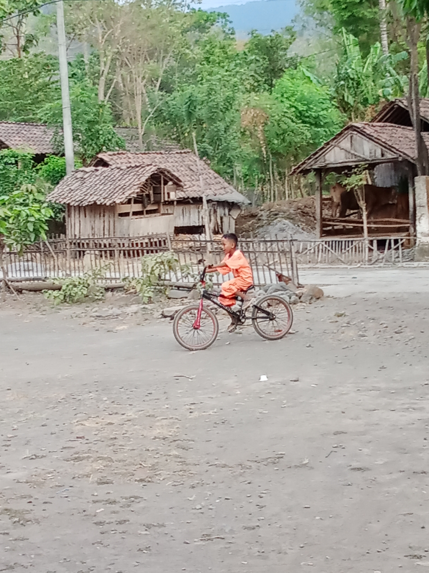 A young boy in an orange jumpsuit is riding a bicycle with red rims on a dirt path. He is in the foreground of the image, with a rural village setting behind him. The village consists of several wooden structures with thatched roofs. In the background, there are trees and a hillside. The overall scene depicts a simple, rural environment.