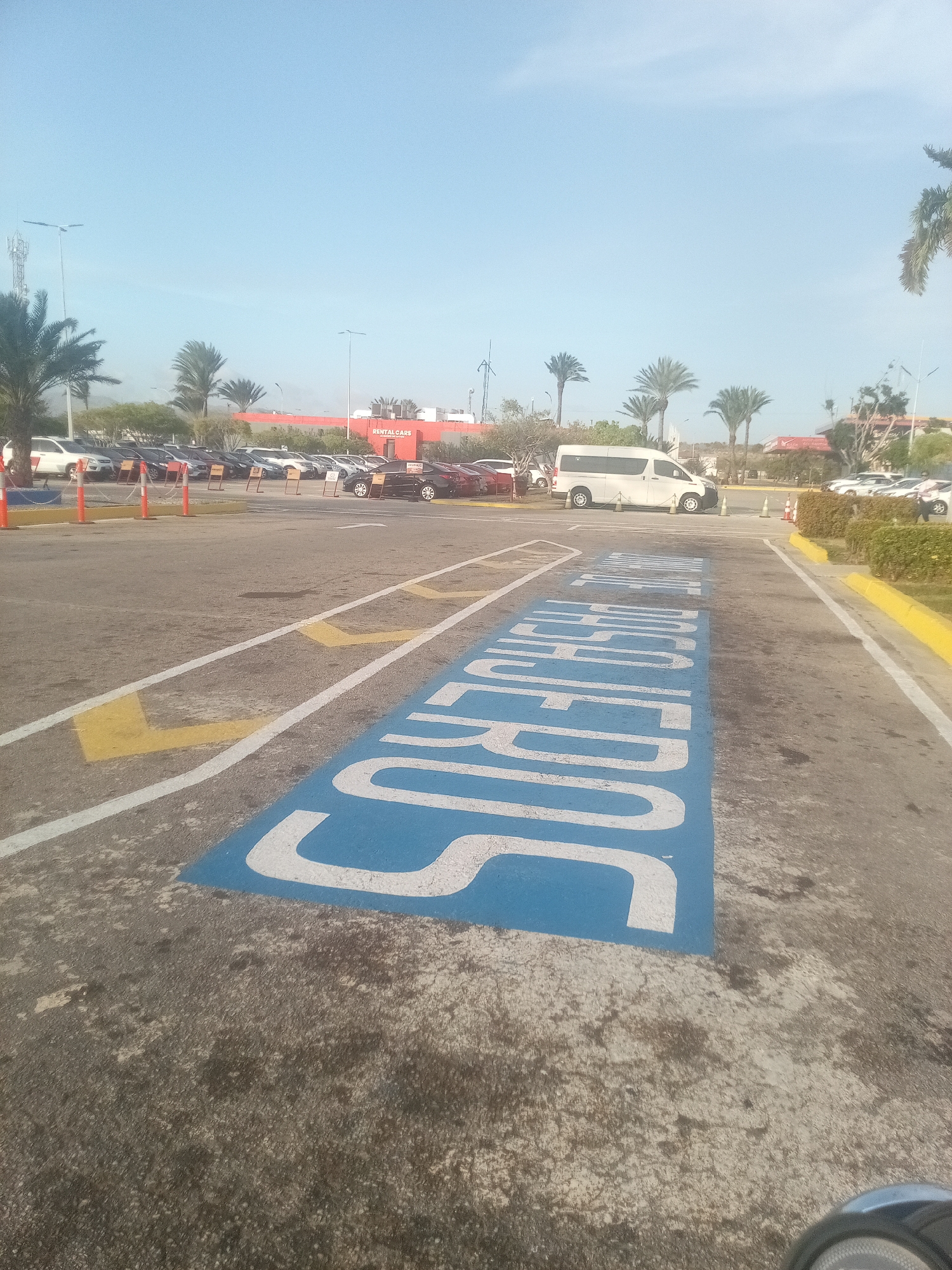 The image shows a parking lot in Cotoperíz, Venezuela. The lot is paved and contains multiple parking spaces, including one specifically marked for "SUPERVISORS" in blue paint. Cars and a white van are parked in the lot. Palm trees are visible in the background, and the sky is clear. A building with the words "RENTAL CARS" is also in the background. Orange traffic cones are placed near some parking spots.