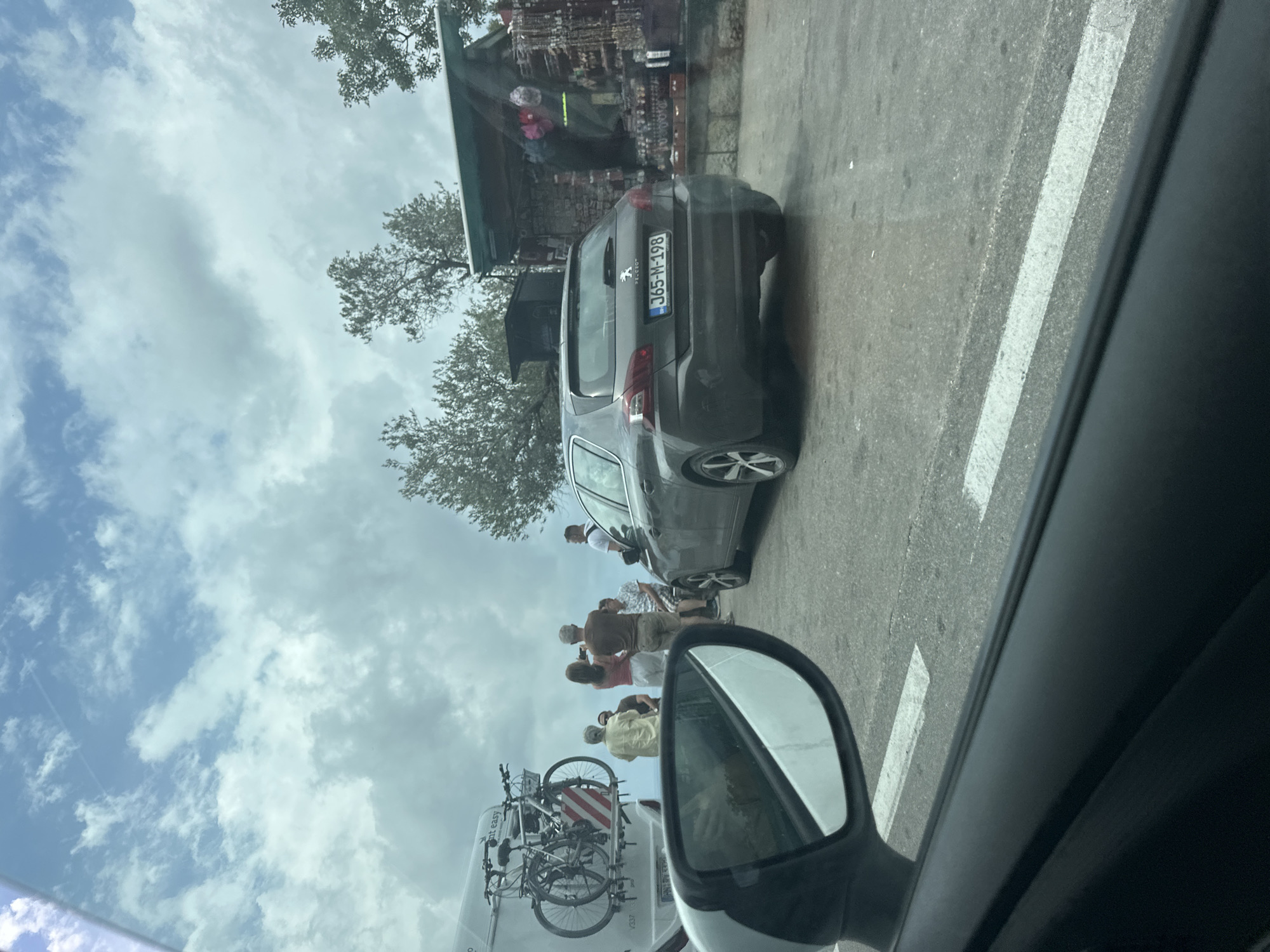 The image, taken from within a vehicle, depicts a casual daytime scene along a roadside in Pržno, Montenegro. The sky above is bright and mostly cloudy.
In the mid-ground, a dark grey Peugeot hatchback is parked on the right side of the road, facing away from the camera. Its Montenegrin license plate, "J65-M-198," with "MNE" visible on a blue strip, is clearly legible. Behind the car, a small, rustic roadside stall or shop is visible, displaying various goods, possibly souvenirs or handicrafts. Trees with sparse foliage frame the upper and middle sections of the scene.
Several people are gathered on the roadside. Some individuals are standing near the parked Peugeot, while others are closer to the left, partially obscured by the viewer's car's side mirror. Through this mirror and in the lower-left corner, part of a white van is visible, equipped with a bicycle rack holding two bicycles. The people appear to be casually dressed, suggesting they might be tourists or locals making a stop.
The foreground shows the dark frame of the viewer's car window and its side mirror. The road surface features distinct white lines marking the edge. The overall impression is a relaxed, perhaps scenic, stop along a route, likely for browsing the roadside stall or enjoying the location.