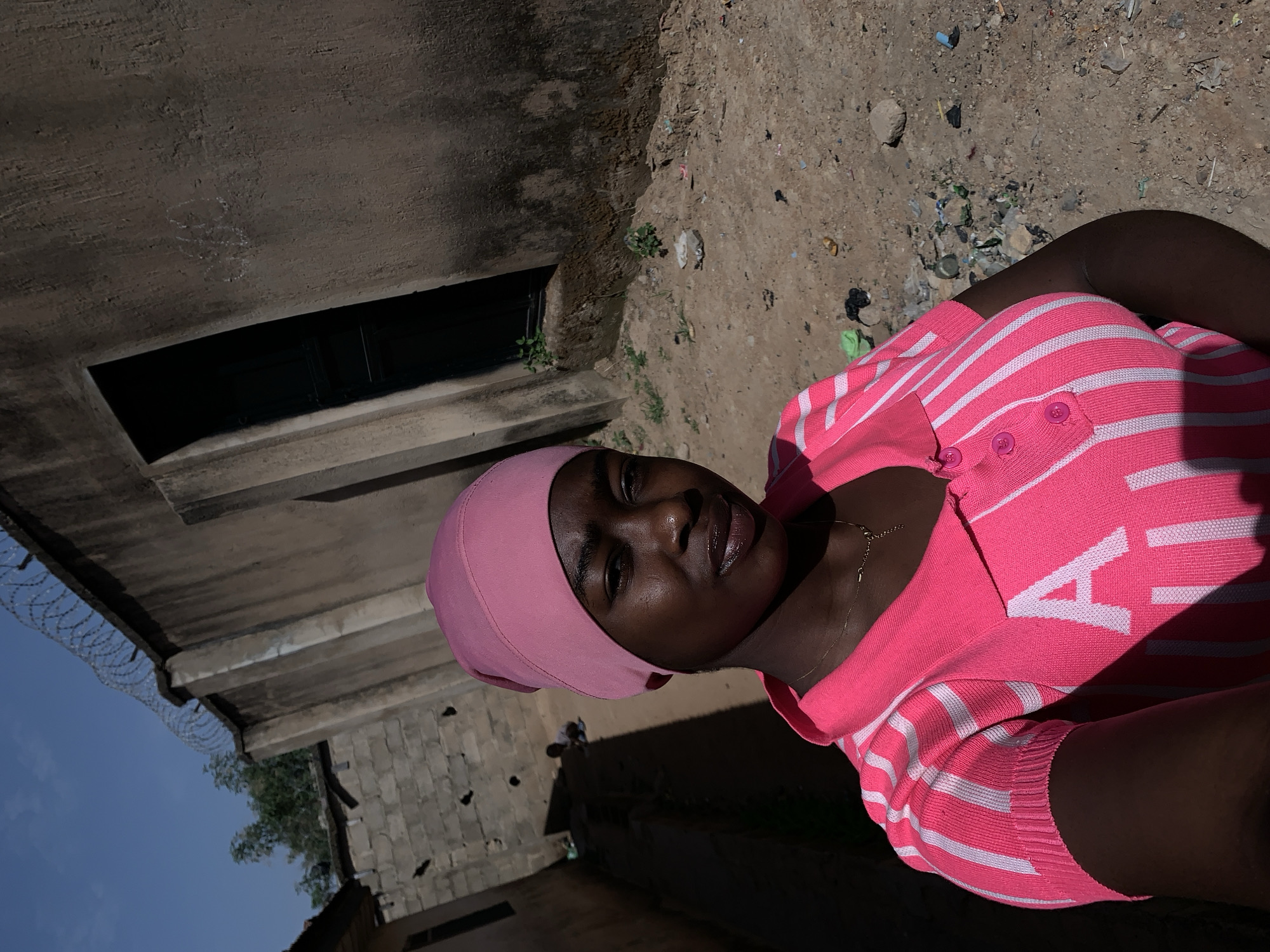 A woman with dark skin wearing a pink head covering and a pink and white striped sweater stands outdoors. The background features a weathered building with a dark window opening. Debris litters the ground beside the woman. In the background are buildings and blue sky. The scene appears to be Rafin Par, Nigeria.