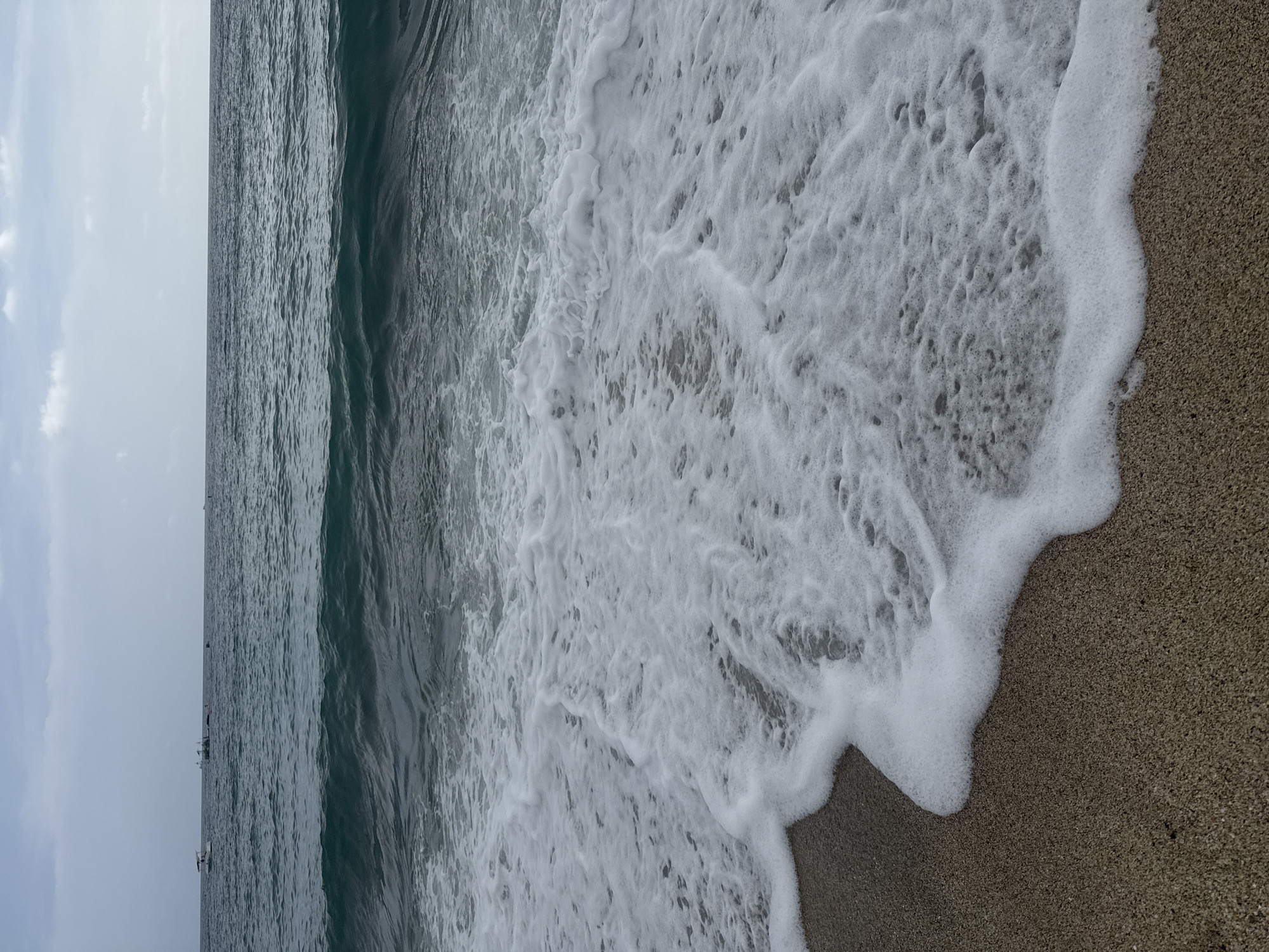 The image shows a beach scene in Alanya, Turkey. The view is from the perspective of someone standing on the beach, looking out at the water. The waves are crashing onto the shore, and the foam is visible. The sand is a light brown color. The sky is overcast. There are a few boats visible in the distance.