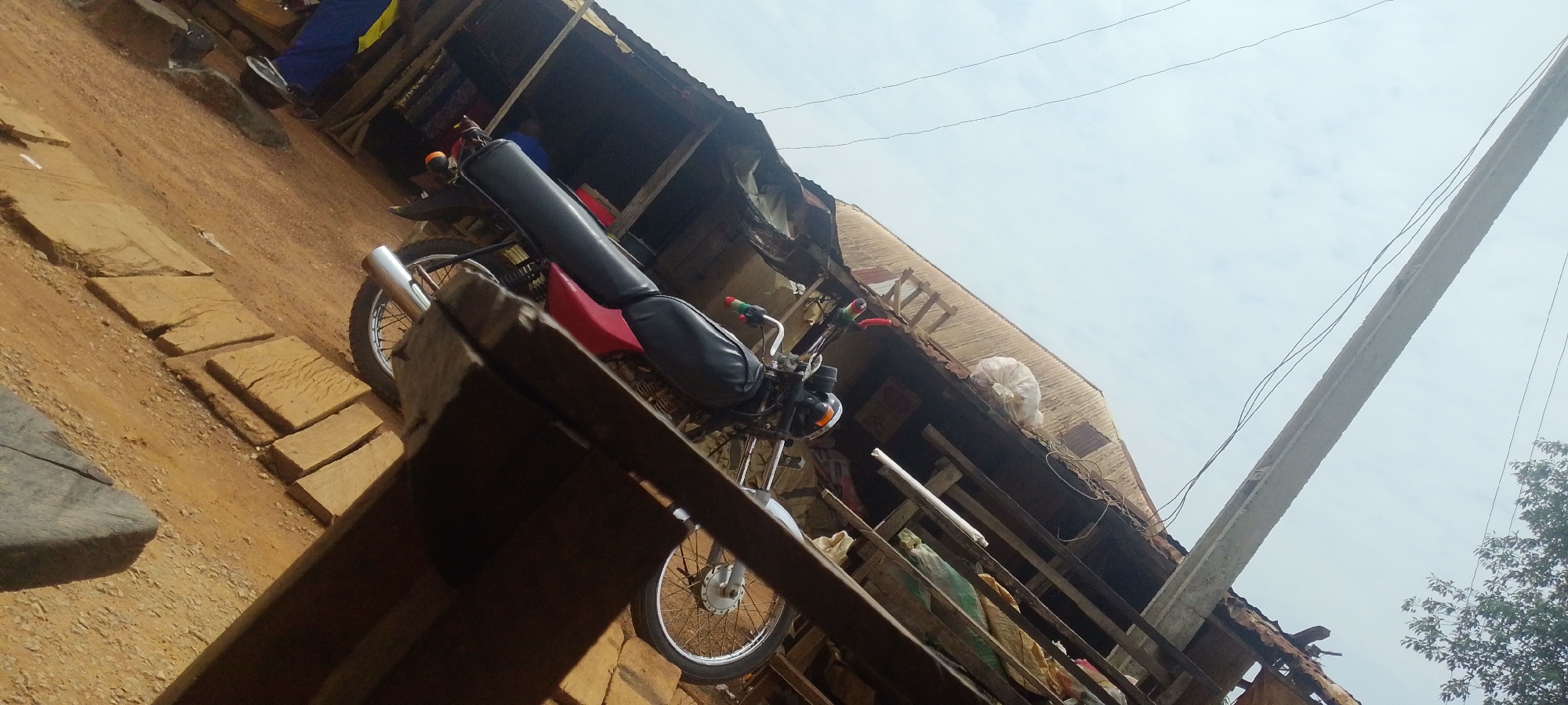 In the city of Baissa, Nigeria, a motorcycle with a long, black padded seat and a red base is parked on a dirt ground with several flat, light brown paving stones laid out in a row. The motorcycle is positioned at an angle, with its handlebars and front wheel visible. Behind the motorcycle, a weathered, open-air structure with a corrugated metal roof is partially visible. The structure appears to be a stall or shop, with some items and debris scattered around. The overall environment suggests a rural or semi-urban setting, characterized by the natural ground, simple construction, and a cloudy sky.