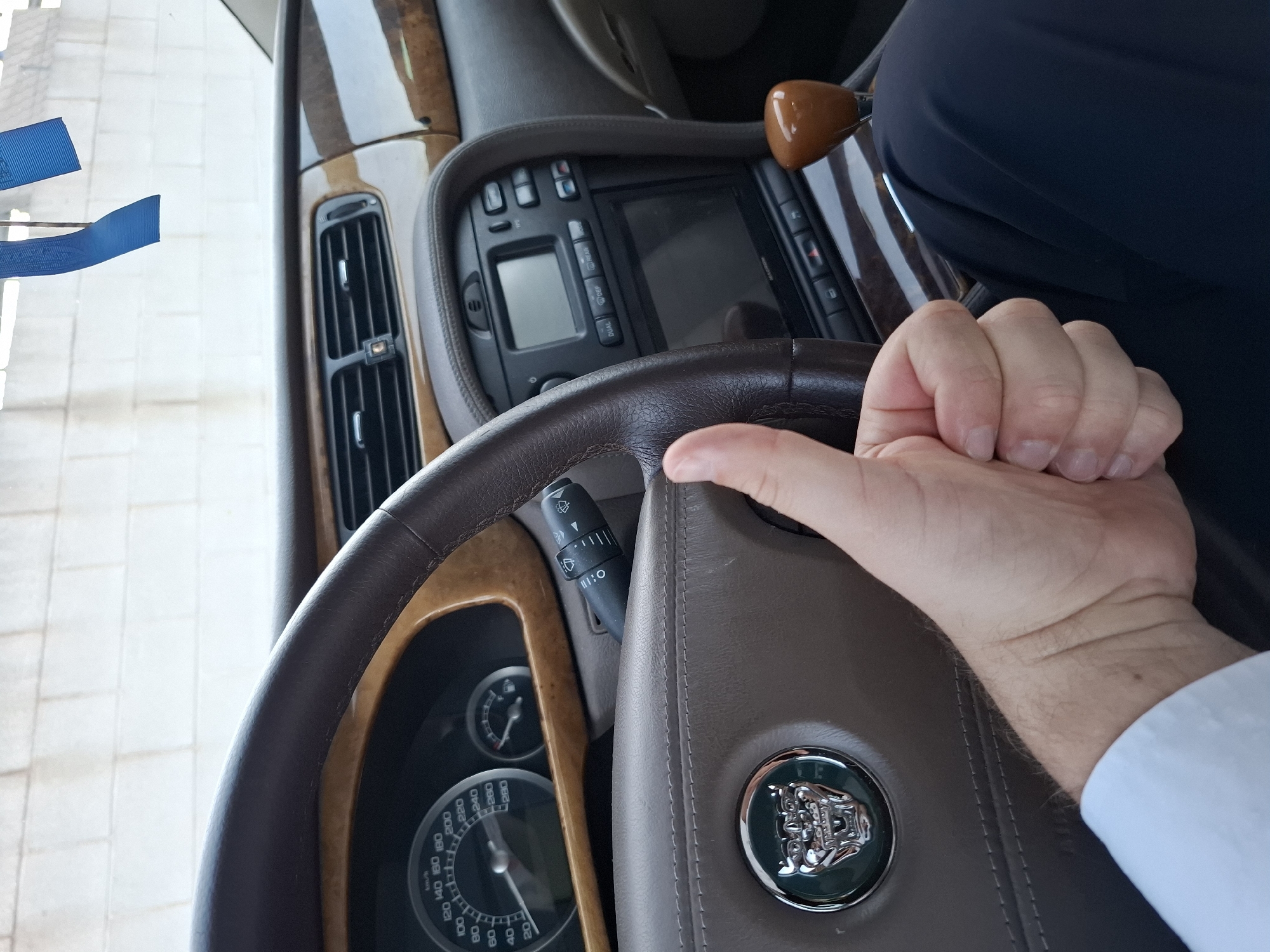 Here is a forensic description of the image:
The image shows the interior of a vehicle, specifically the steering wheel of a Jaguar. The steering wheel is brown leather with a central emblem. The dashboard is visible in the background, featuring a wood trim, air vents, and a display screen. The gauge cluster can also be seen, showing the speedometer, marked in km/h. A hand is resting on the steering wheel, wearing a white shirt. The background also shows a glimpse of the exterior with white tiled pavement. The image does not include enough information to determine if the image was taken in Quart de Poblet, Spain.