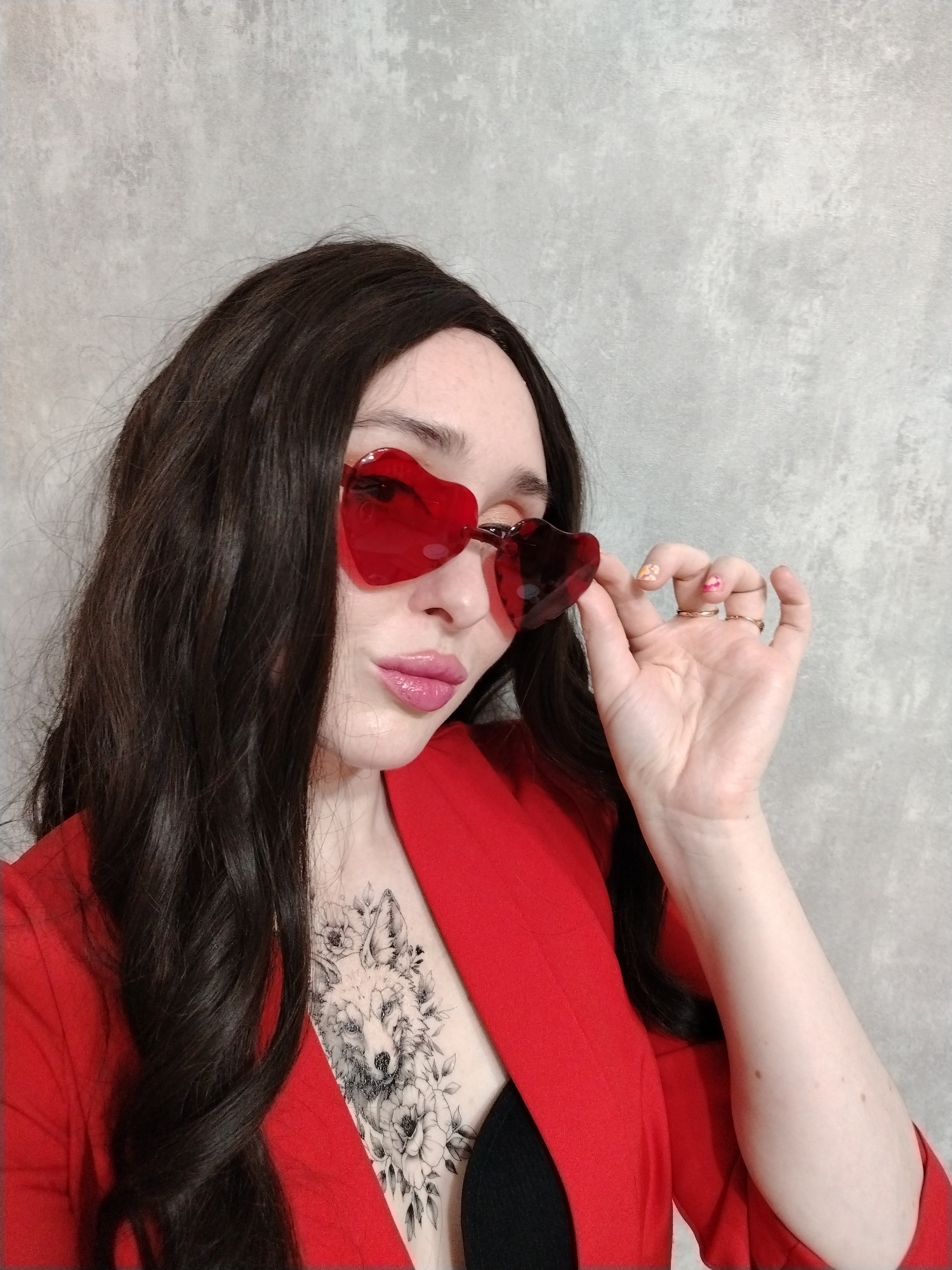 A young woman poses indoors against a mottled gray wall. She is wearing a long, dark, wavy wig and red heart-shaped sunglasses. Her lips are glossy and pink. She is wearing a red blazer, exposing a bit of a black top underneath. A black-and-white wolf tattoo is visible on her upper chest. Her right hand is raised, holding the frame of the sunglasses. She wears two simple gold rings and her nails are painted with varied designs. The location in the city of Izhevsk, Russia is indeterminable based on visual information.