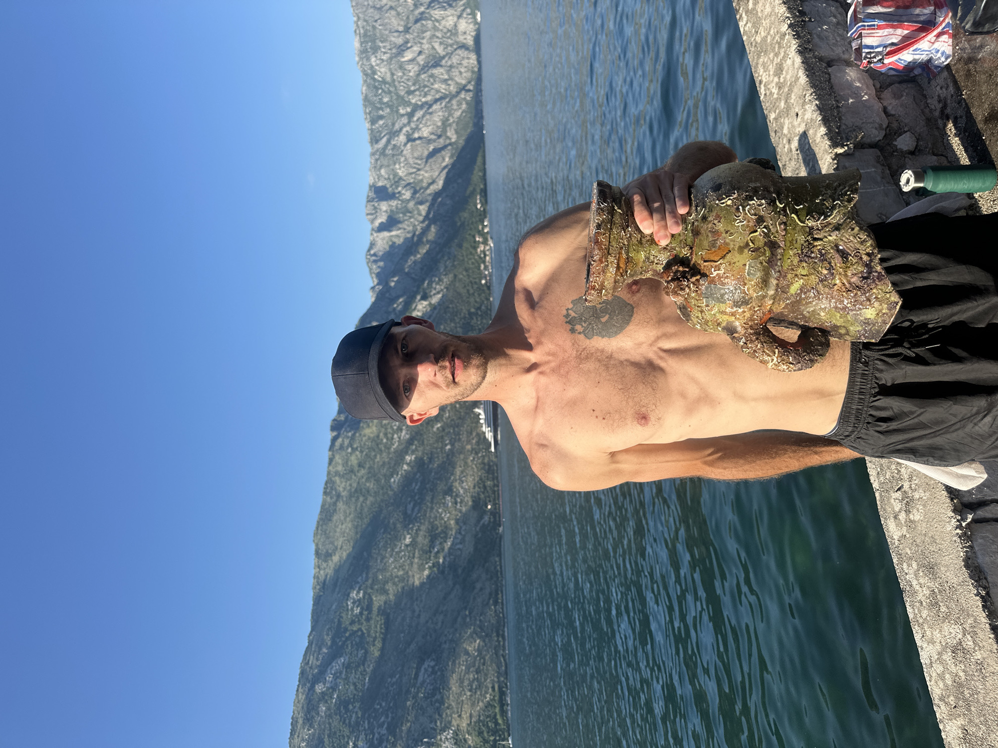 An image taken outdoors in Prčanj, Montenegro, depicts a male figure standing beside a body of water with a mountainous background under a clear blue sky.
The central figure is an adult male, appearing to be in his late 20s to early 30s, with fair skin and short facial stubble. He wears a black baseball cap and black athletic shorts with a visible grey waistband and white drawstring; he is shirtless. A tattoo resembling an anatomical heart with root-like elements is visible on his left pectoral. He holds a large, irregularly shaped object, heavily encrusted with marine growth, predominantly dark green, brown, and off-white. The object appears to be a relic, possibly a vase or urn, with a wide body, a narrower neck, and two arm-like protrusions, all covered in barnacles and other marine organisms. His gaze is directed forward, towards the camera, with a neutral expression.
The man stands on a light-grey concrete or stone seawall, constructed from stacked blocks. To his right, on the wall, a striped bag (red, white, and blue) and a small, dark green cylindrical bottle or container are present. A white cloth or towel is partially visible, tucked into his shorts on his right side.
The background features a large, calm body of dark blue-green water, indicative of a bay or fjord. Steep, rocky mountains, partially covered in sparse vegetation, rise directly from the water on both the left and right sides of the frame. In the distance, a large white cruise ship or yacht is visible on the water towards the center-right, and a smaller sailboat is present further left. The sky is uniformly clear and bright blue, suggesting fair weather and strong sunlight, casting visible shadows on the man's face and the object he holds.