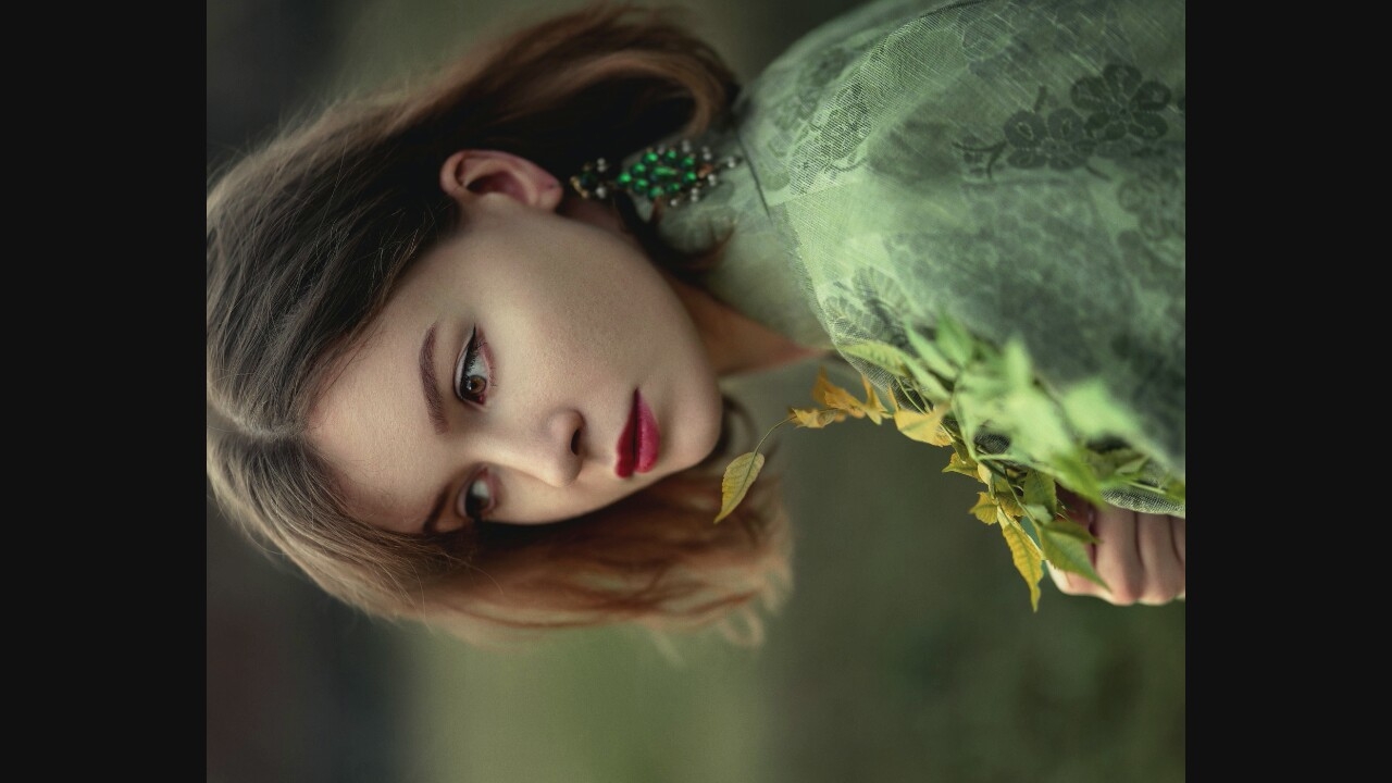 A young woman with fair skin and auburn hair is shown in a medium shot, looking to her right. She is wearing a green, textured garment with floral patterns and a large, ornate green earring on her right ear. She holds a sprig of yellow-leaved branches in her left hand, which rests on her chest. The background is a blurred expanse of muted greens, suggesting an outdoor, natural setting, possibly a field or park, though no specific urban landmarks are visible. The lighting is soft and diffused.
