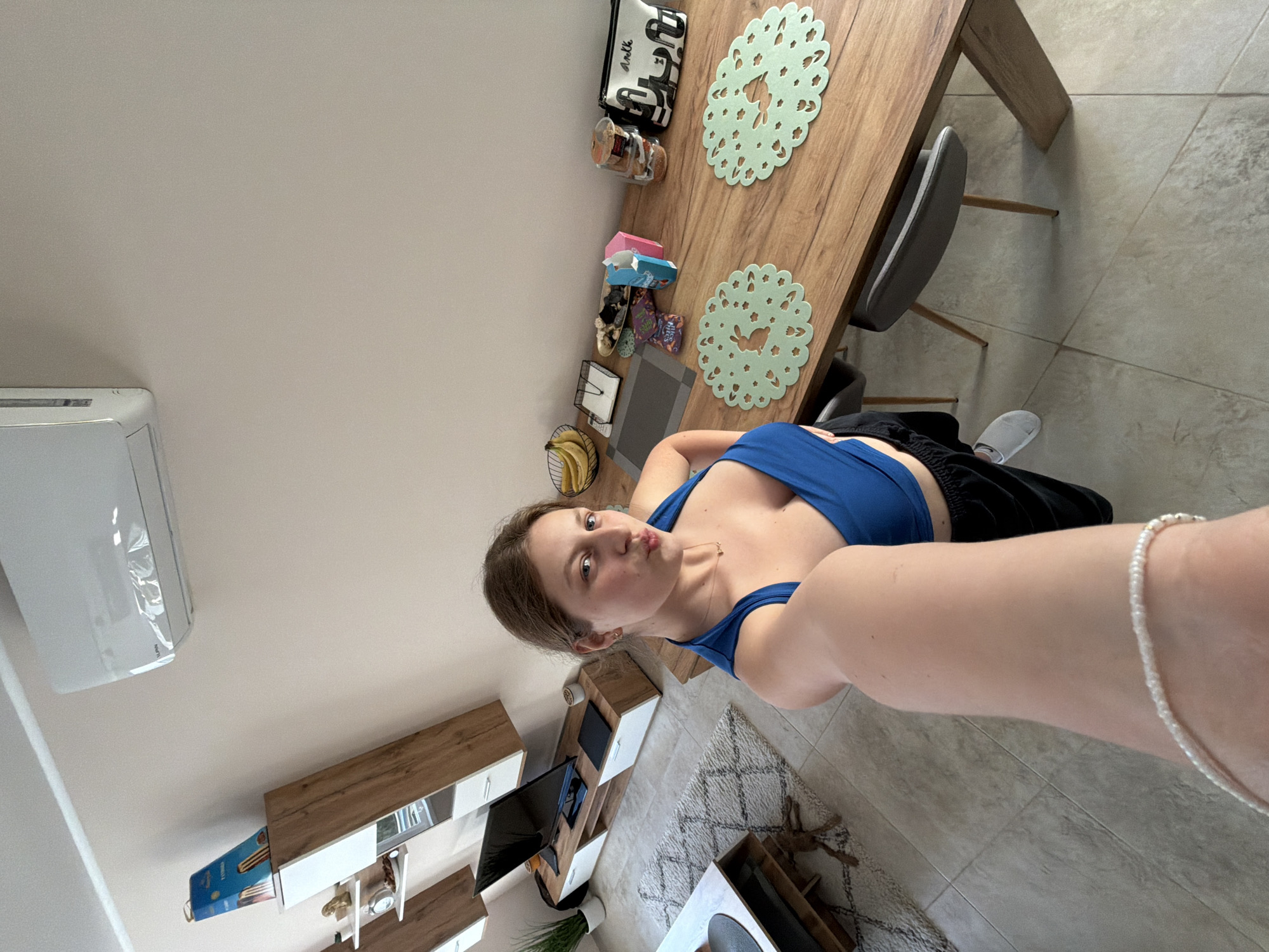 A young woman in a blue top and black shorts is taking a selfie in what appears to be a living room and dining area. The room has light-colored walls and tile flooring. Visible in the background are a wall-mounted air conditioner, a low-profile entertainment center with a TV, and a rug. A dining table is situated nearby, adorned with decorative placemats and various items. The location cannot be determined as Litakovo, Bulgaria from the provided information.