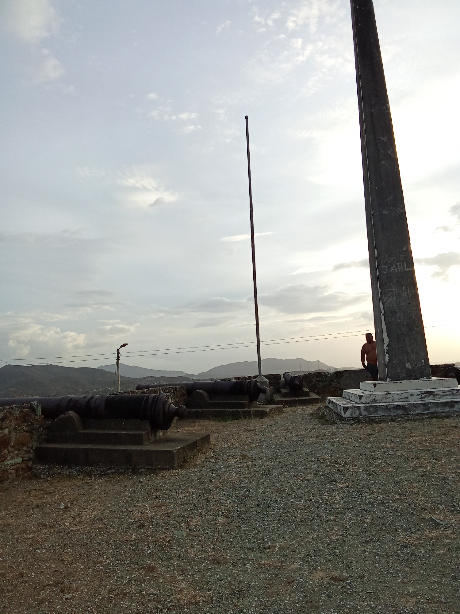 This image shows a stone wall overlooking a mountainous landscape with a cloudy sky. Four cannons are positioned along the wall. A tall stone obelisk stands to the right of the cannons. A person is standing near the obelisk. The ground in the foreground is covered with gravel. A flagpole stands between the cannons and the obelisk. This scene is in the city of Juan Griego, Venezuela.