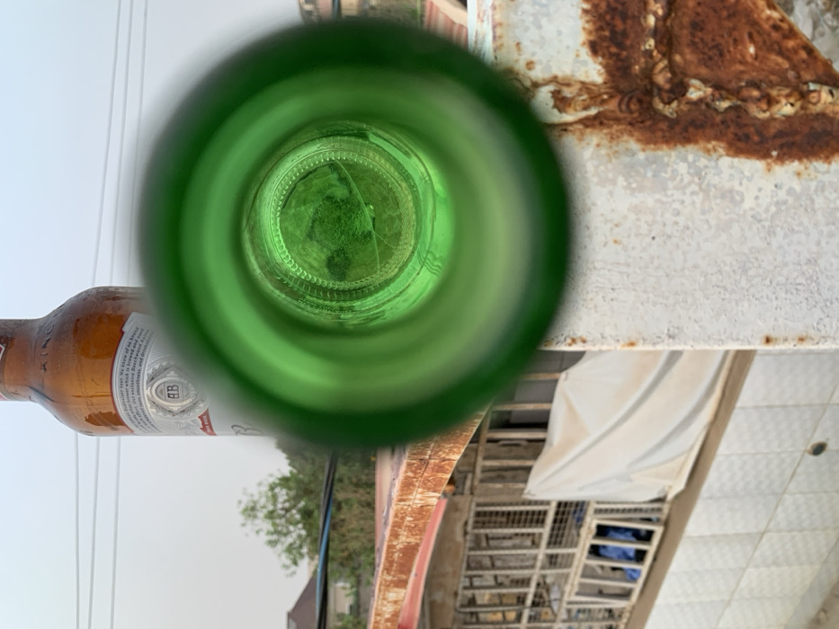 The image depicts a view through the neck of a green bottle, possibly a beer bottle, capturing an outdoor scene. A portion of a brown bottle is visible on the left. The interior view through the green bottle neck shows greenery, possibly foliage. The surrounding environment includes a white tiled surface, a white covering, metal structures, and an aged metal surface with rust. The sky is visible in the distance, suggesting an outdoor setting.