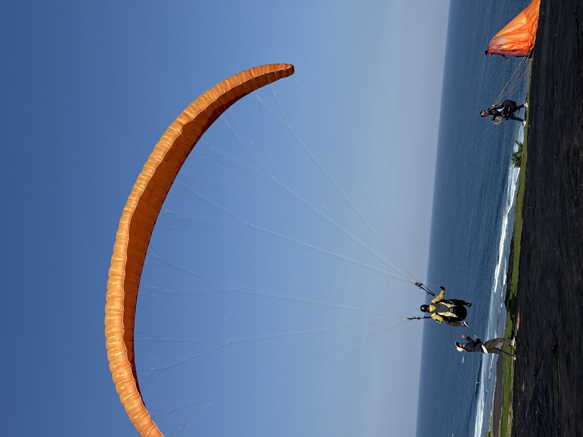In Kuta Selatan, Indonesia, on a bright, clear day, a paraglider is captured mid-air against a vast blue sky. The paraglider's wing, a vibrant orange, arches prominently across the frame. Beneath the wing, a person in a yellow jacket and black pants is suspended in a harness, their face obscured by a helmet. Below and to the left of the airborne paraglider, another individual wearing a white hat, dark jacket, and grey pants stands on a dark, textured ground, looking up at the paraglider. To the right, on the same dark ground, another person in dark attire is handling a large, bright orange paraglider wing that is spread out on the ground, with lines extending from it. In the background, the deep blue ocean meets a white-capped shoreline, with a strip of green land bordering the dark ground in the foreground.