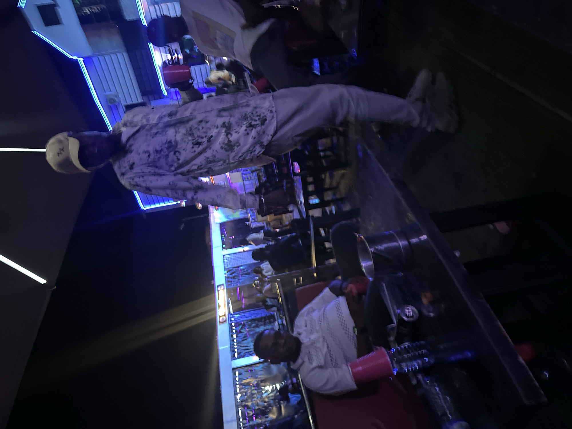 Here's a description of the image:
The image appears to be taken indoors, likely at an entertainment venue. A man wearing a patterned shirt, light-colored pants, and a baseball cap is standing near game machines. Another individual is seated at one of the game machines, holding a pink cup. Blue and purple lighting is visible in the background, suggesting the presence of screens or decorative lights. The location may be Tumfure, Nigeria, but there is no explicit evidence within the image to confirm it.
