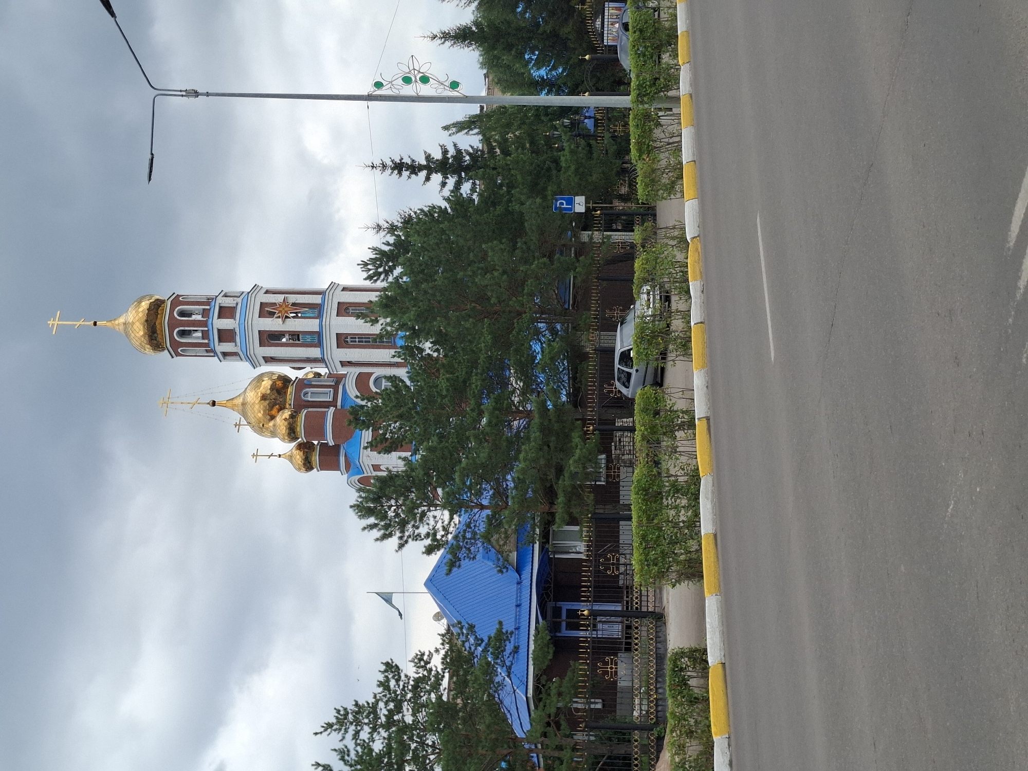 The image shows a street view in the city of Petropavl, Kazakhstan. In the background, a large church with golden domes and a tall bell tower dominates the skyline. The church is red brick with blue accents and white trim. In front of the church, there is a smaller building with a blue roof, surrounded by a decorative black and gold fence and manicured green hedges. Several pine trees are also visible. A street with a grey asphalt surface runs through the foreground, bordered by a curb painted with alternating white and yellow stripes. There are a few cars parked along the side of the street behind the fence. The sky is overcast and cloudy.