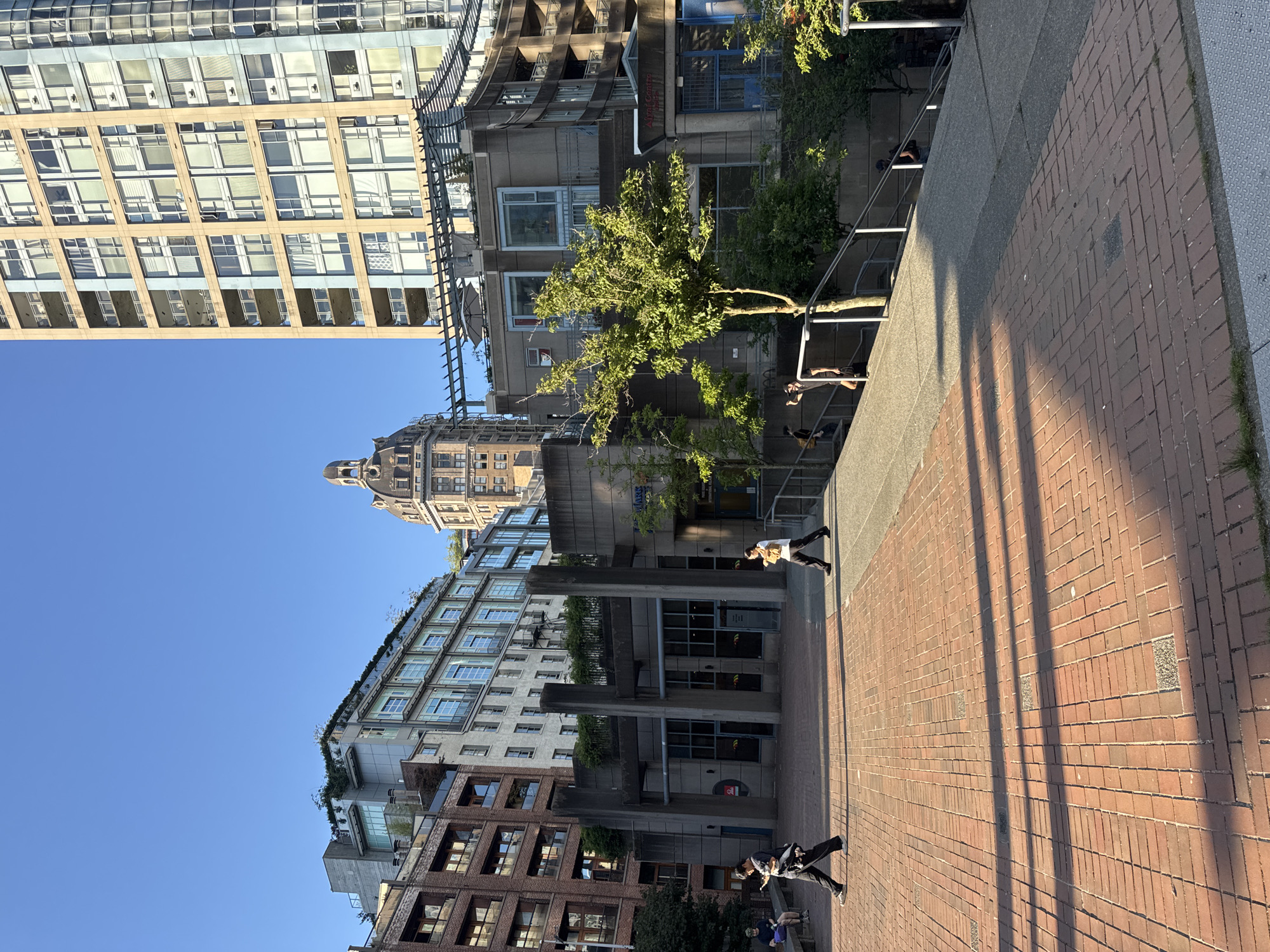 This image depicts an outdoor plaza in Vancouver, Canada, likely on a sunny day with a clear blue sky. The plaza is paved with red bricks, laid in a herringbone pattern, with some square concrete inlays. Long shadows are cast across the plaza, indicating the time of day is likely morning or late afternoon.
In the mid-ground, a group of people are present. Two individuals are walking across the plaza from left to right. One person, wearing a dark shirt and pants, is walking with a phone in their hand and what appears to be a black bag slung over their shoulder. Another person, wearing a white shirt and dark pants, is walking towards the right. Further back, several other figures are seen walking and standing, including a person sitting on a bench in the far left of the frame.
The background is dominated by several multi-story buildings of varying architectural styles. To the left, there is a brick building with numerous windows, followed by a lighter colored building with a more modern design and greenery on its rooftop. In the center, a distinctive building with a domed roof stands out. To the right, a tall, modern high-rise building with a facade of light-colored panels and blue accents rises into the sky. Between these buildings, there are other structures with concrete facades, balconies, and windows. Trees with green foliage are also visible, some growing from planters on the buildings. A metal trellis structure is visible extending from one of the buildings. Staircases and railings are also present, suggesting the plaza is on a slight incline. Signs are visible on some of the buildings, but are not legible enough to read.
