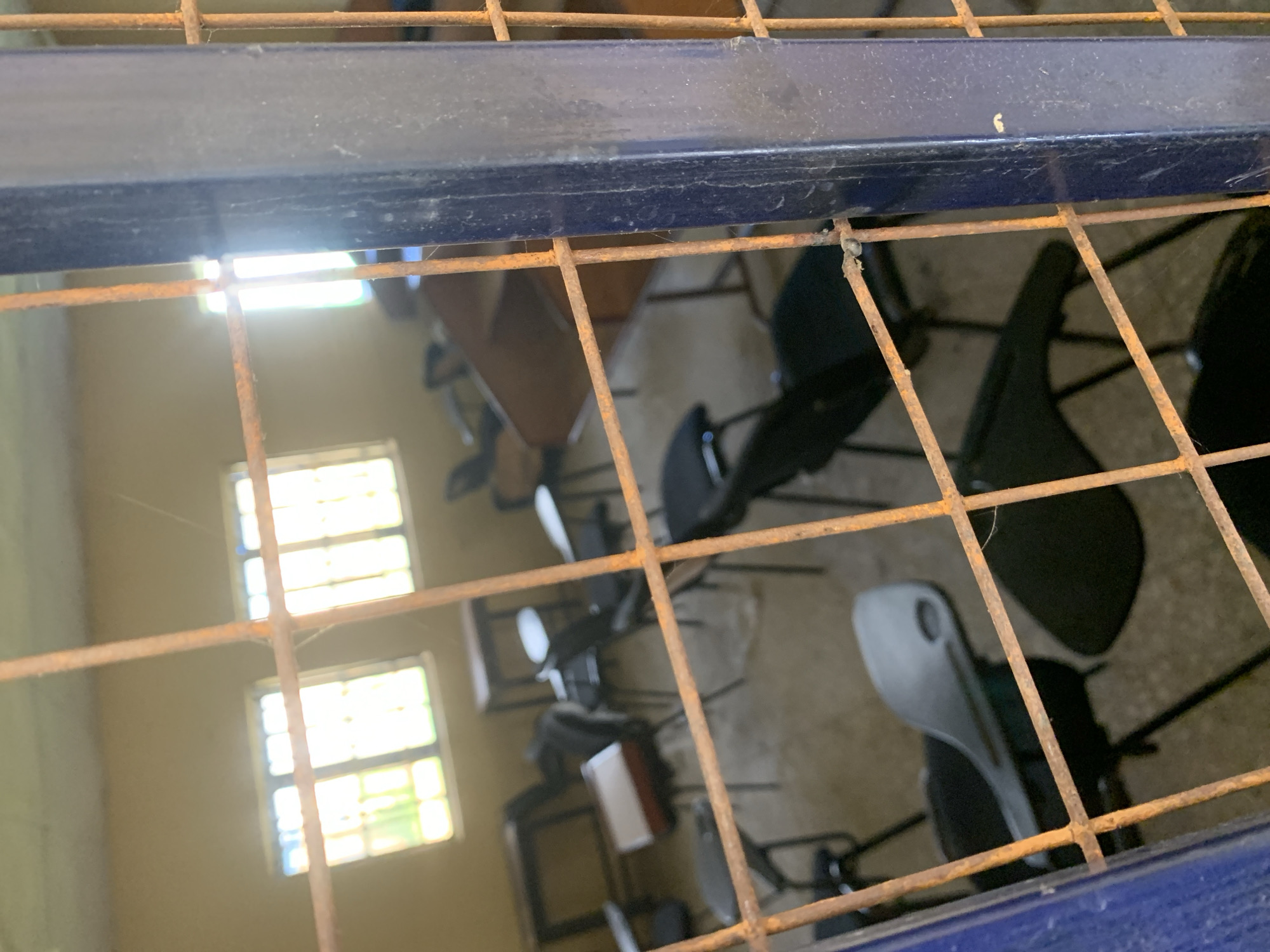 An interior view of an empty classroom in Jalingo, Nigeria, is seen through a rusty metal grate. The room appears to be abandoned or unoccupied, with several chairs and desks scattered around. Sunlight streams through two windows, illuminating dust particles in the air. The floor is concrete and appears dirty. There are no people in the image. The overall impression is one of disuse and neglect.