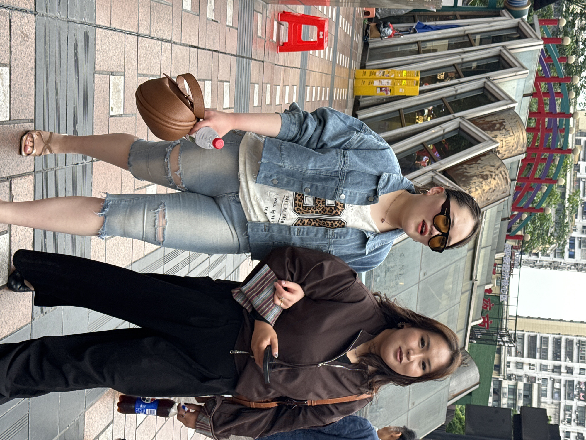 Two women are standing next to each other outdoors in Yuzhong District, China. The woman on top is wearing denim shorts, a white t-shirt, and a blue denim jacket; she has a brown purse and is holding a water bottle. The woman below her is wearing black pants, a brown jacket, and has a crossbody bag; she is holding a smartphone in one hand and a bottle of soda in the other. They appear to be on a paved walkway in front of a building with unique architectural details. Tall buildings are visible in the background.