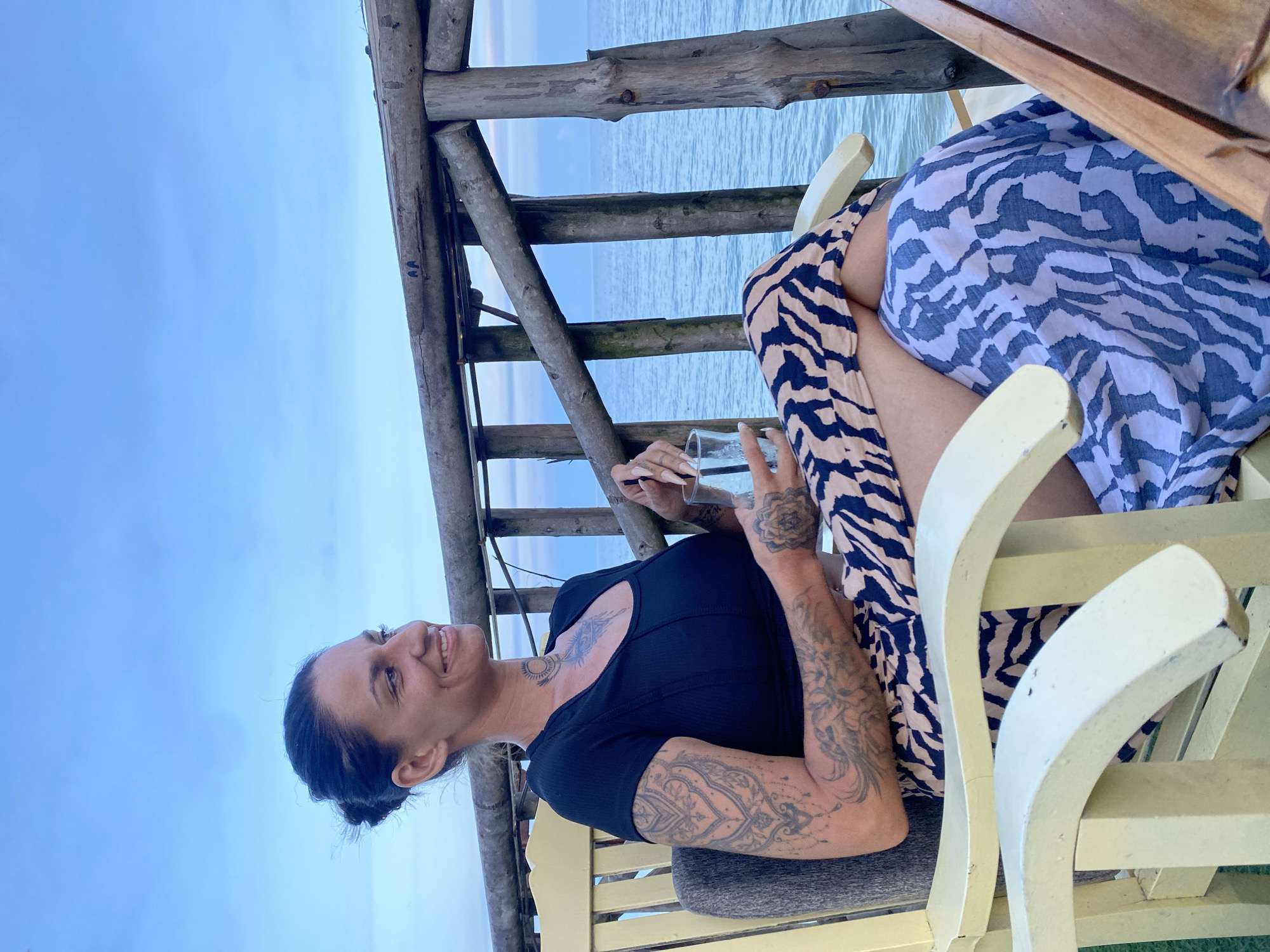 In Phu Thuy Ward, Vietnam, a woman with dark hair tied back sits on a light-colored wooden chair, facing left and looking out at the ocean. She is wearing a black t-shirt and a patterned skirt with beige and dark blue zebra-like stripes, with a blue and white patterned fabric draped over her lap. The woman has extensive tattoos on her left arm, chest, and neck. She is holding a clear glass in her right hand, which appears to contain a clear liquid. The background features a rustic wooden railing and the calm blue ocean under a slightly overcast sky. The image is taken from a medium distance, capturing the woman from the chest up, with emphasis on her pose and facial expression.