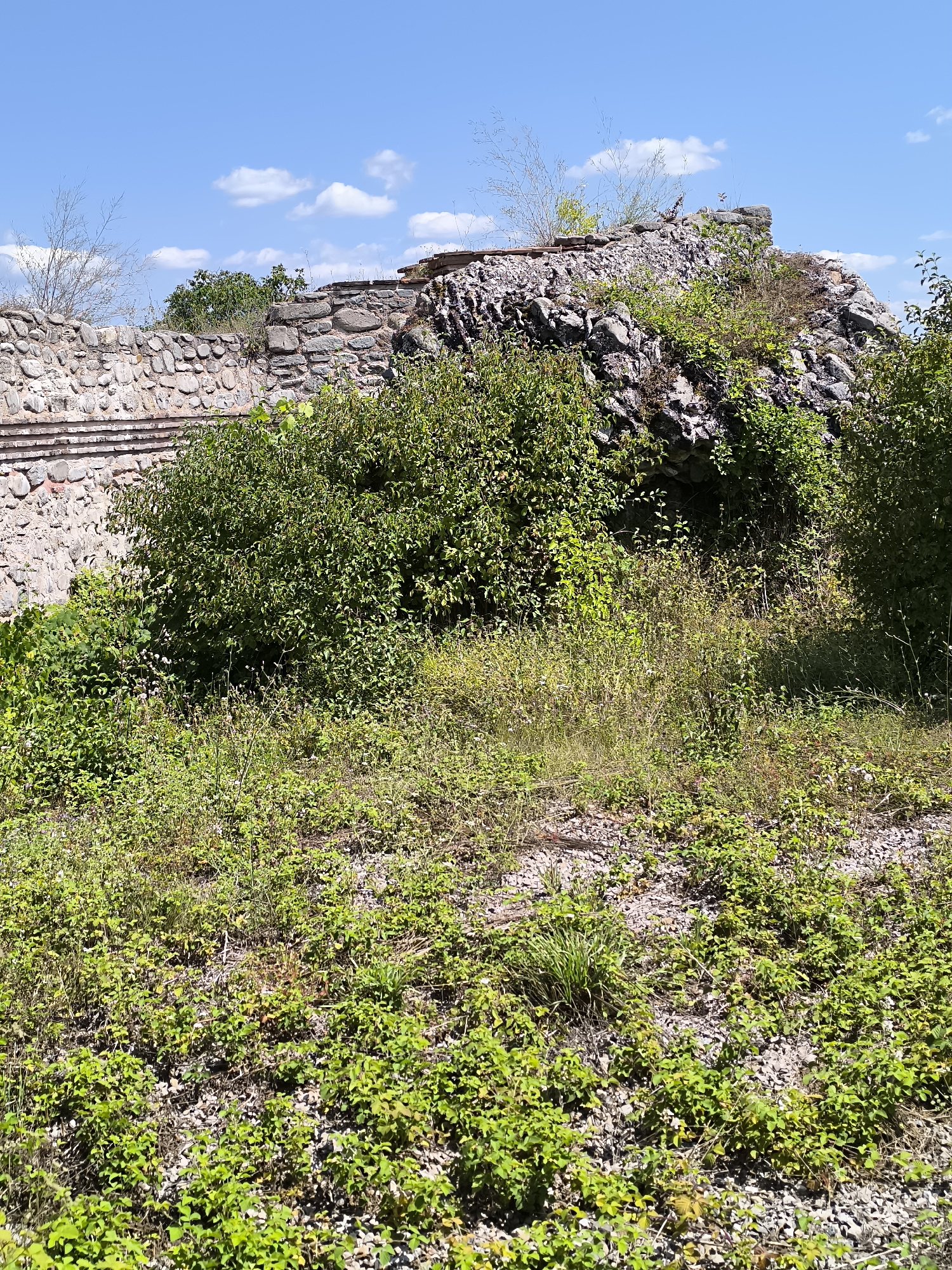The image shows the remains of an ancient Roman fortress in Garmen, Bulgaria. The fortress is made of stone and is partially overgrown with vegetation. The sky is blue with some clouds. The fortress is located on a hill overlooking the surrounding landscape.