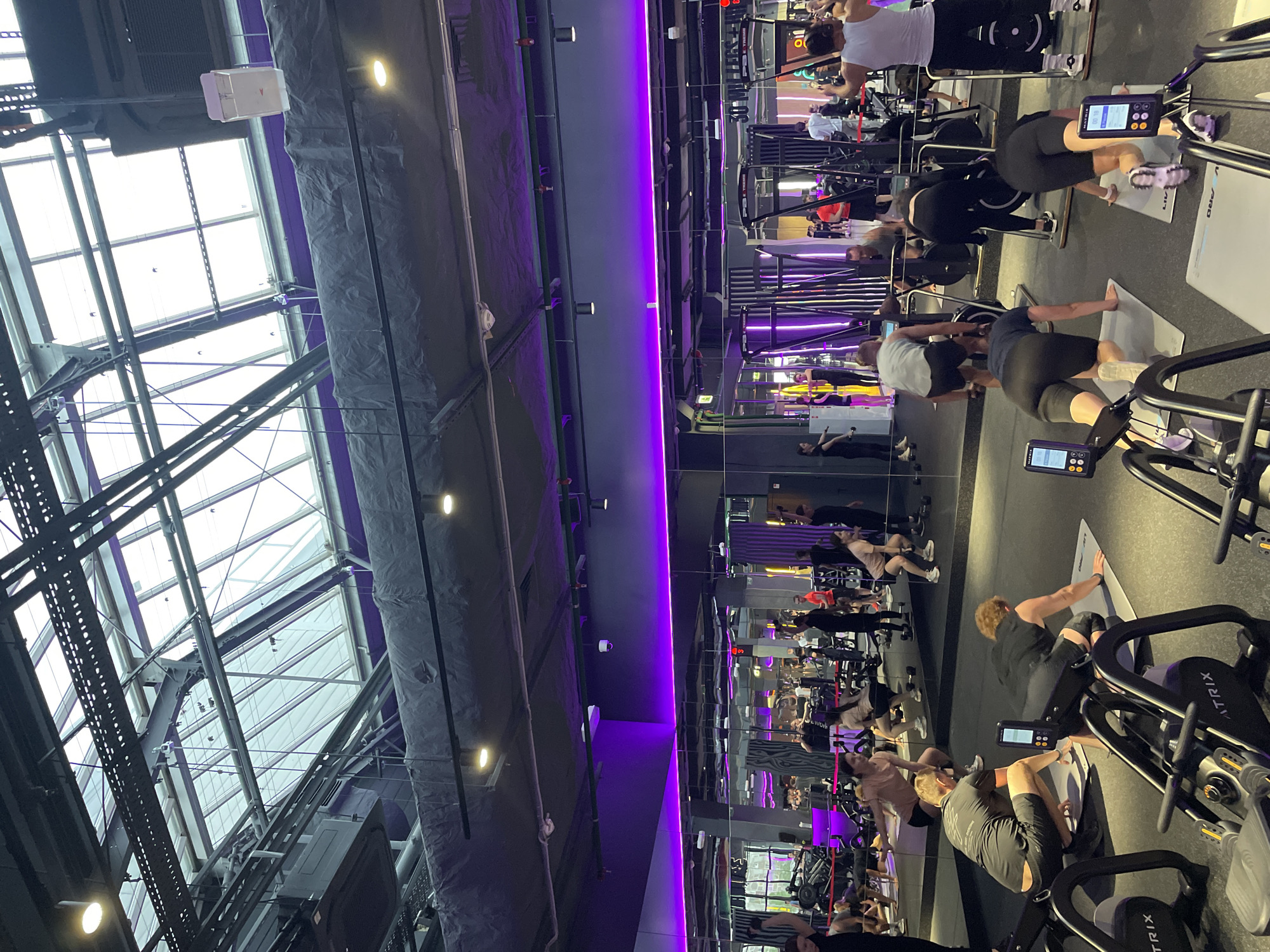 The image shows a gym in Kazan, Russia. It is daytime, as light streams through the large, geometric, steel-framed windows. Numerous individuals are exercising on rowing machines. The interior is modern, with a mirrored wall reflecting the exercise area, and a dark gray ceiling adorned with purple lighting. The rowing machines are positioned on mats.