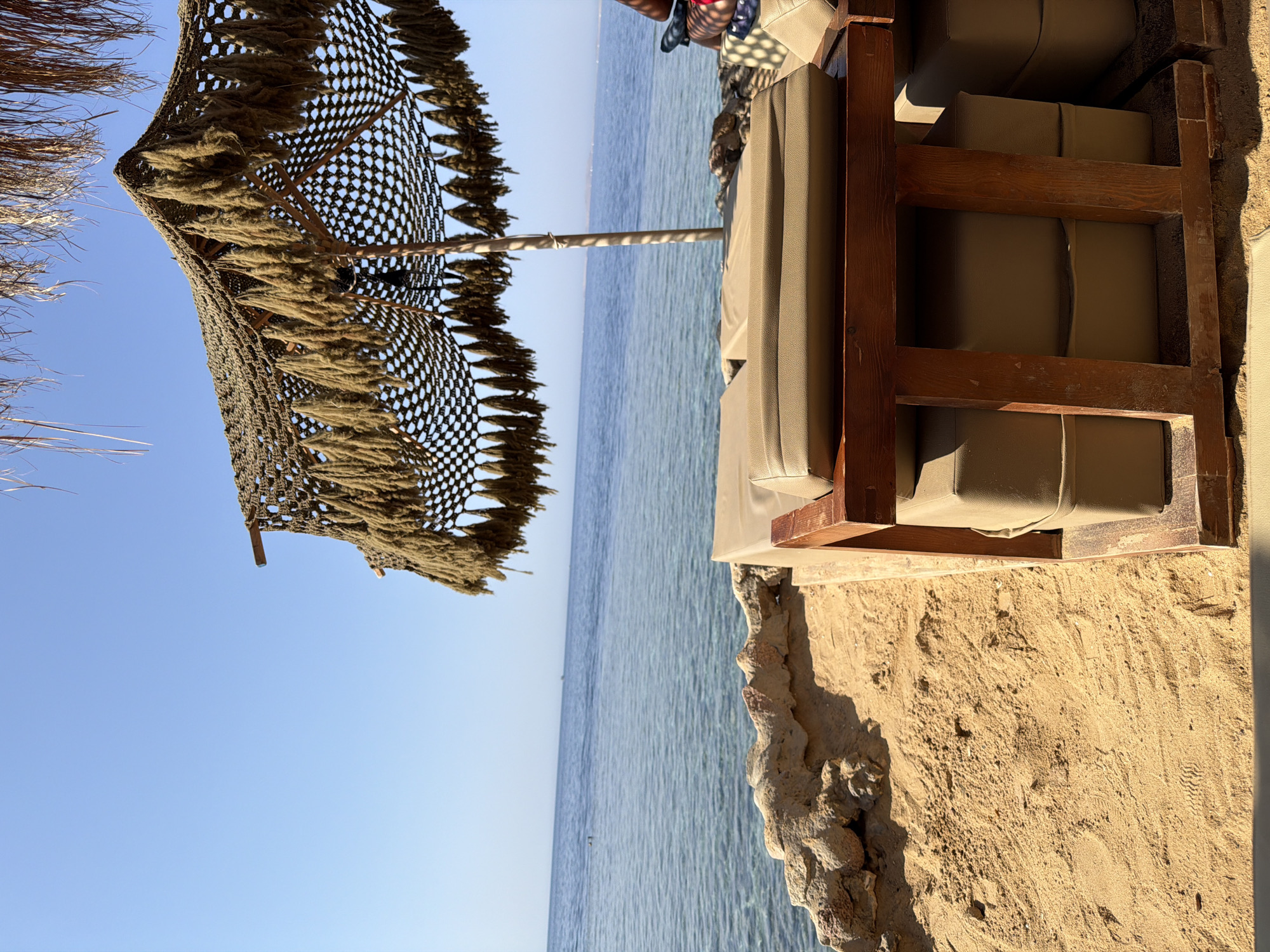 The image depicts a serene beach scene on a sunny day, likely in Sharm El Sheikh, Egypt. In the foreground, a rustic wooden lounge chair with plush, beige cushions is set on a sandy beach. The chair is positioned to offer a view of the calm, blue sea. To the left of the chair, a low, rocky wall separates the sand from the water.
Above the lounge chair, a unique umbrella provides shade. The umbrella's canopy is made of netting with numerous thick, rope-like strands hanging down, creating a textured fringe. A thin, white umbrella pole supports the structure. In the distance, the vast expanse of the sea meets a clear, light blue sky. The water appears tranquil, with subtle ripples and a hint of clarity revealing the seabed near the shore.
The overall atmosphere is one of relaxation and leisure, typical of a beach resort. The natural elements of sand, sea, and sky are complemented by the simple, comfortable furniture and the distinctive umbrella, suggesting a well-appointed beachfront.