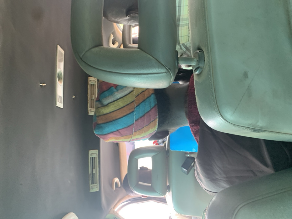 The scene depicts the interior of a vehicle, likely a passenger van or bus, in Alizaga, Nigeria, viewed from a rear seat looking forward. Three individuals are visible from behind. In the immediate midground, a person wears a distinctive multi-colored striped head covering in shades of pink, orange, yellow, green, and blue, over a dark reddish-brown garment. To their left, another individual wears a dark garment, holding a bright blue object, possibly a folder or tablet, and is seated in front of a light-colored headrest with a rectangular cutout. To the right of the central figure, a third person's head and shoulder are partially visible, wearing a light-colored plaid shirt. The vehicle's interior features a dark grey ceiling with overhead air conditioning vents, including two elongated rectangular vents and a central circular vent panel. The seats are light-colored, possibly beige or light green, showing signs of wear and minor discoloration. Natural light enters from the left side, indicating daylight outside.