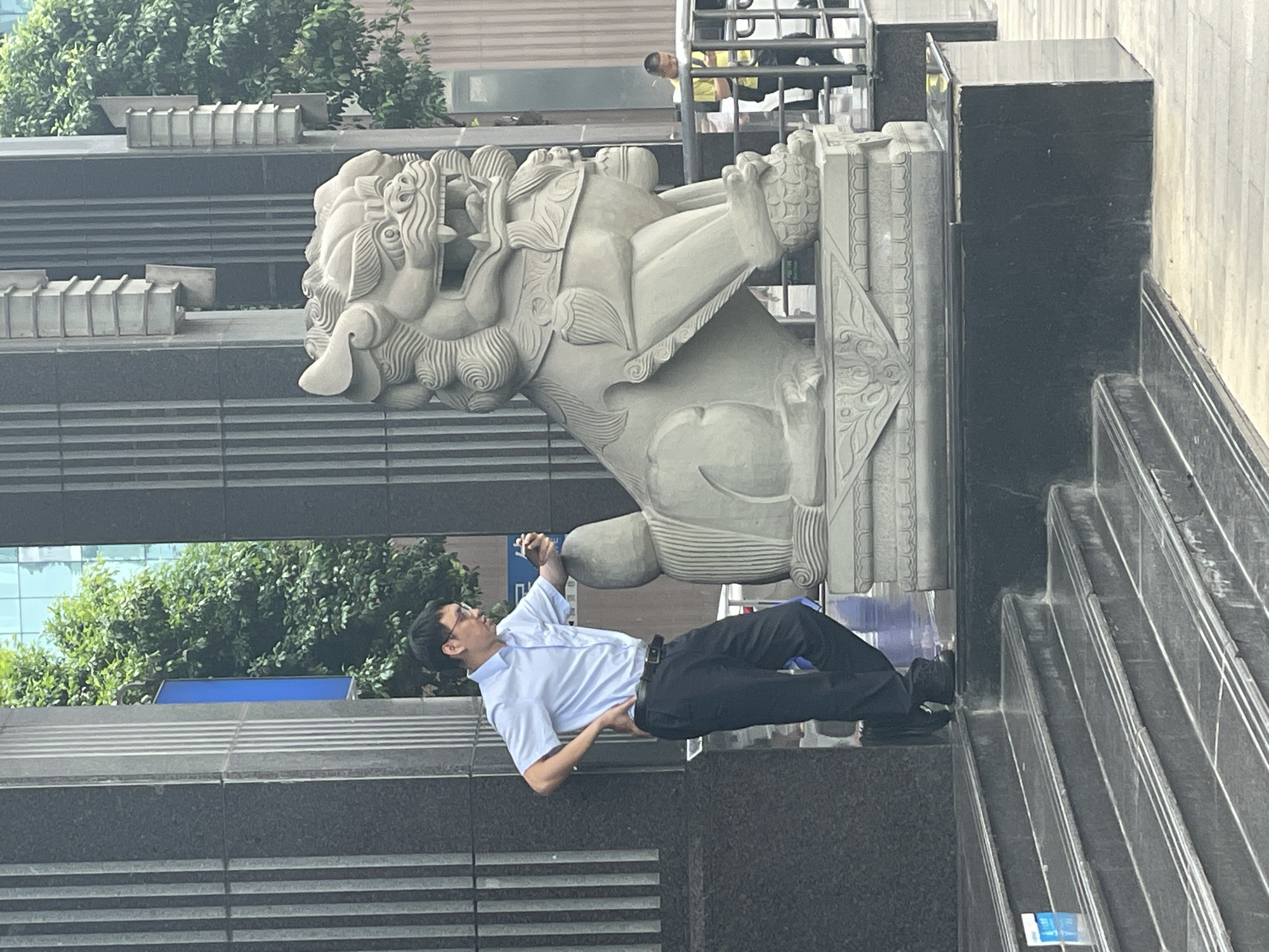 A man in a light blue, short-sleeved shirt and dark pants is interacting with a stone lion statue in what appears to be the Yuzhong District, China. The statue is positioned on a platform adjacent to a set of stairs. The man is looking up, possibly taking a photograph or observing the statue closely. The area is urban, with trees and a multi-story building visible in the background. The statue and man appear to be near the entrance or exterior of a building.