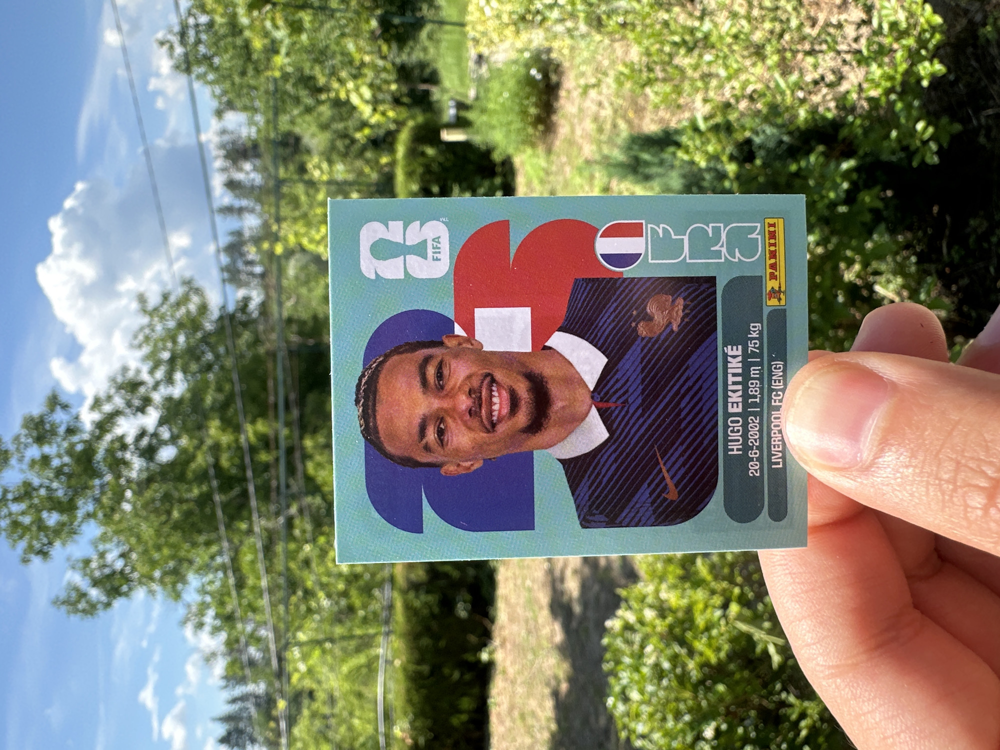 An extreme close-up outdoor shot shows a hand holding a soccer trading card featuring Hugo Ekitiké, a French footballer. The card displays his image, name, birthdate (20-6-2002), height (1.89 m), weight (75 kg), and club (Liverpool FC (ENG)). The background is blurred, but suggests a natural environment with trees, grass, and a bright sky with clouds. The location is identified as Prisovo, Bulgaria.