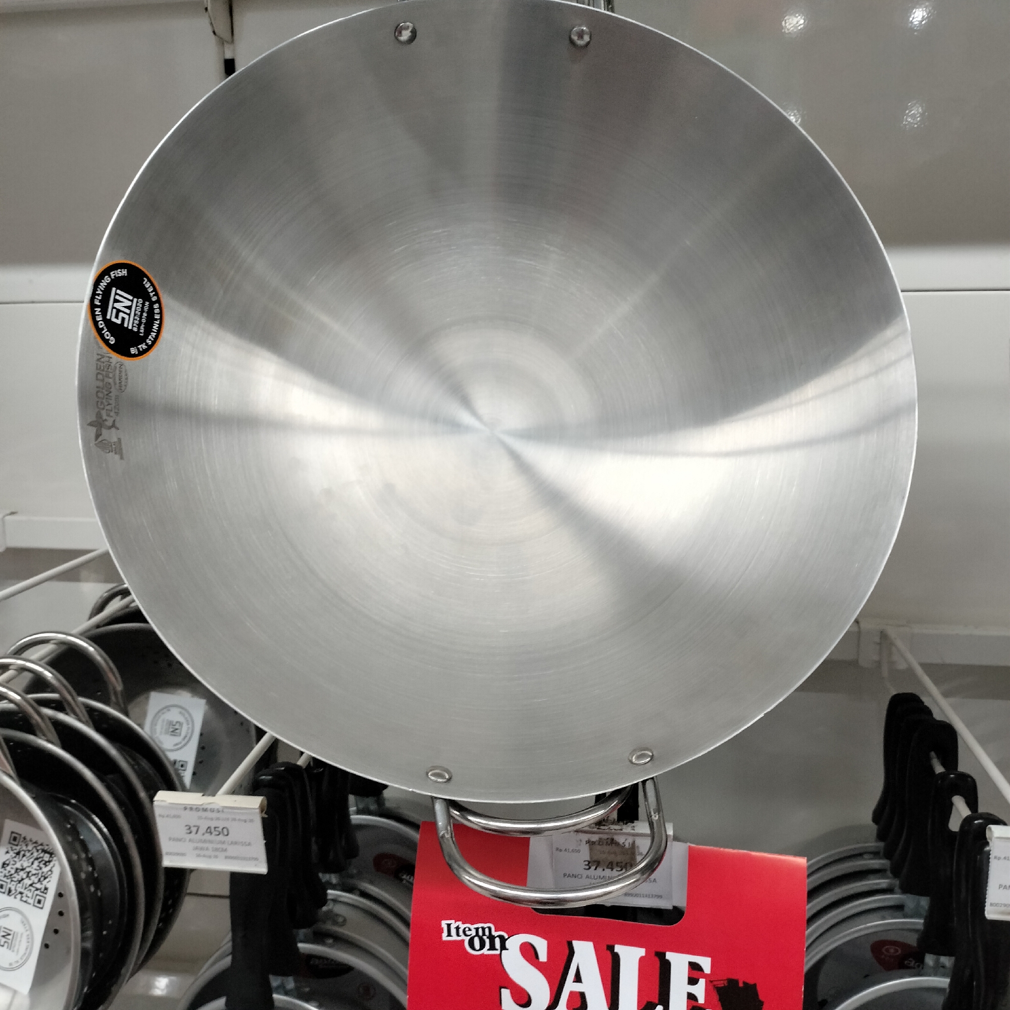 The image displays a close-up, overhead view of a large, circular, brushed metal pan, likely a wok or a frying pan, with two metal handles attached to its rim. The pan's surface exhibits concentric circular striations characteristic of brushed metal. A circular sticker with orange and black lettering is adhered to the left side of the pan. The sticker contains text such as "SNI," "GOLDEN FLYING FISH," and "STAINLESS STEEL."
Below the pan, other kitchenware, including stacked circular pans with black handles, are visible. A red sign with the words "Item on SALE" printed in large white letters is positioned in the lower right quadrant of the image. Several white price tags are attached to the items. One visible price tag displays "37,450" and "PANO ALUMINIUM LARISSA."
The setting appears to be a retail environment, possibly a kitchenware store, with shelves and hanging displays. The lighting suggests an indoor setting with overhead illumination. There are no people or animals present in the image. The background consists of muted, neutral-toned shelving and walls, typical of a commercial display.