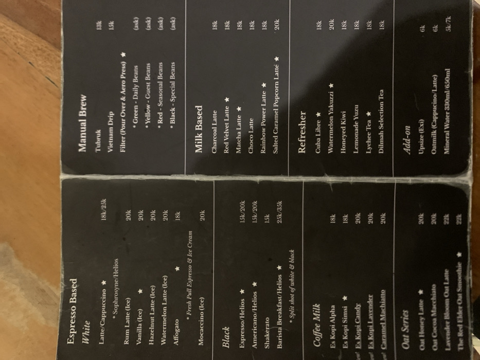 The image is a close-up view of a laminated menu board, likely at a coffee shop. It shows a list of coffee drinks, with prices in Indonesian Rupiah. The menu is divided into sections, including "Espresso Based," "Manual Brew," "Milk Based," "Refresher," "Oat Series," and "Add-on." The prices are listed in the 10,000s, 10k, 15k, 20k, 25k, 35k. Some drinks have stars next to them. The menu is displayed on a brown surface, likely a table or counter. The location indicated by the coordinates (-7.751, 113.213) is in the city of Probolinggo, Indonesia. The image is well-lit and appears to be taken during the day.