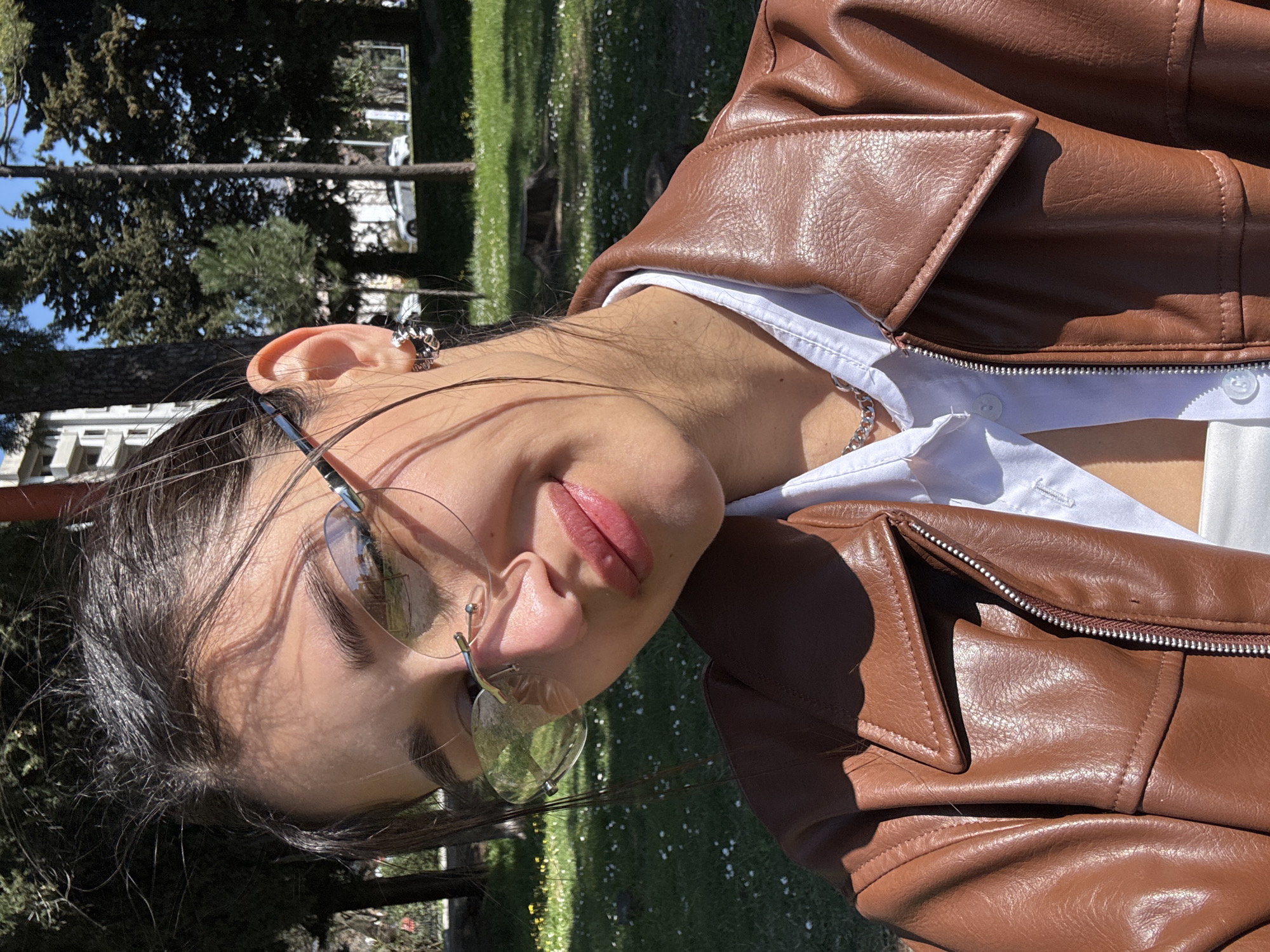 A young woman is outdoors on a sunny day in Bar, Montenegro. She is wearing glasses, a white collared shirt, a silver necklace, a brown leather jacket, and a silver earring. The background consists of green grass and trees. The woman's face is prominent, and she appears to be looking slightly downward. The foreground includes details of her clothing, such as the zipper and pockets of the leather jacket. In the background, glimpses of buildings suggest an urban park setting within the city.