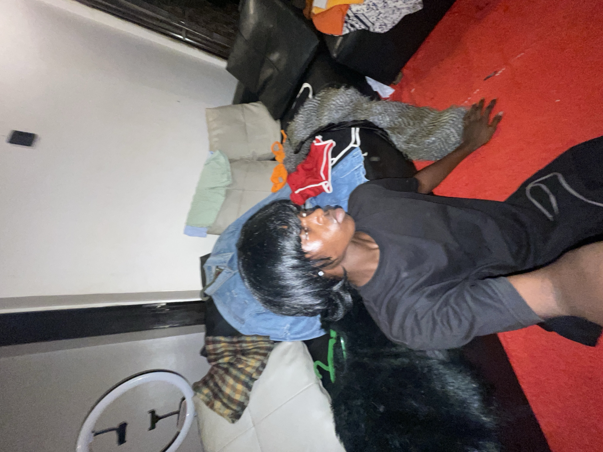 This image captures an indoor scene, possibly a living room or bedroom, depicting a person lying down amidst various household items and scattered clothing. The main subject is a person, likely female, with dark skin and dark, straight hair, wearing a black long-sleeved top. She is positioned on a light-colored couch, with her head angled upwards and her right arm extended onto a vibrant red rug or carpet that covers a significant part of the floor.
The room appears somewhat untidy with clothes strewn about. Piled on and around a black leather couch are several garments, including a light blue denim jacket, a plaid shirt, a red top, an orange top, a light green item, and a grey/patterned fuzzy garment. A white ring light is visible in the lower-left corner of the frame, resting on a surface next to the couch. In the background, a white wall features a small black rectangular object and a dark-colored rail or ledge running horizontally. The lighting is artificial, illuminating the scene and creating some highlights on the person's face. The person's expression appears neutral or relaxed. No visible text is present in the image.