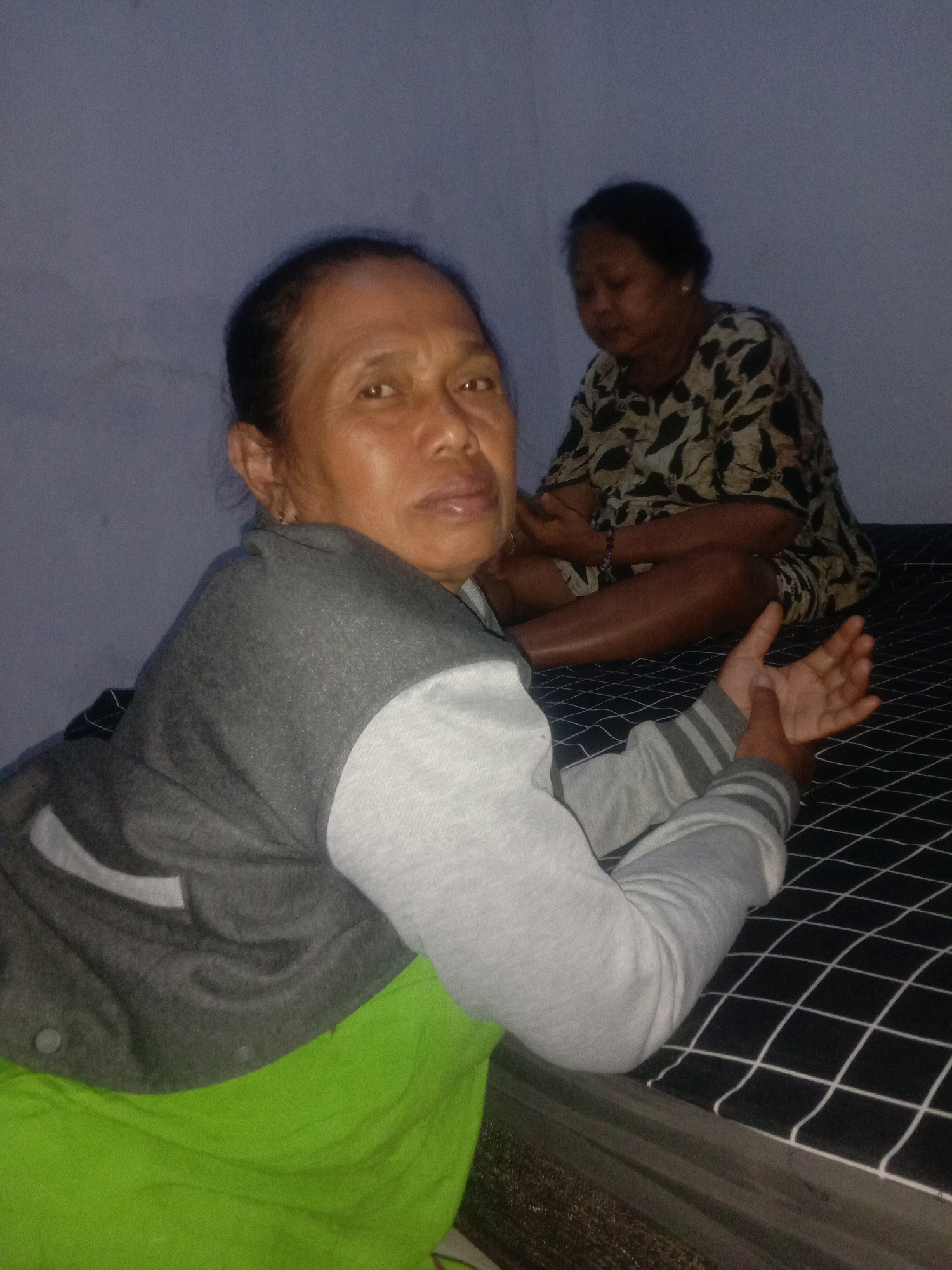 Two women are in a dimly lit room. The woman in the foreground is leaning on her right arm, looking towards the camera. She has brown skin, dark hair pulled back, and is wearing a grey and white jacket over a bright green shirt. The woman in the background is sitting cross-legged on a black and white checkered mattress. She has brown skin and dark hair and is wearing a patterned top. The room appears to be simple, with a plain wall and the mattress possibly being a bed. The location is the city of Tongaswetan, Indonesia.