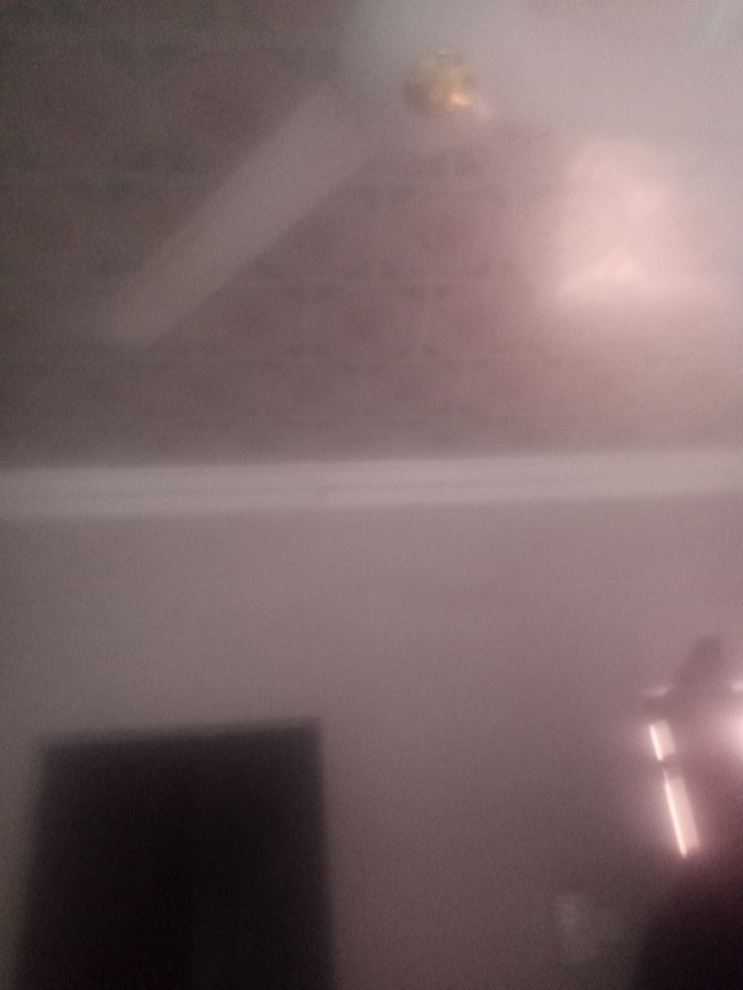 The media file depicts an interior scene, likely a room, with indistinct objects and lighting. The overall impression is of a low-light environment, possibly at night or with artificial illumination.
In the upper portion of the image, a blurred, circular object with a yellowish hue is visible, suggesting it might be a light fixture or a decorative element on a ceiling. A faint, diagonal line across this area could indicate a ceiling fan or a beam of light.
The middle section of the image shows a horizontal, bright band of light, which could be a light source reflecting off a surface or a strip of lighting. Below this, a dark, rectangular shape dominates the lower left quadrant, possibly representing furniture or an appliance.
To the right, in the lower portion, is another bright, elongated light source, possibly a lamp or a screen. The overall color palette is muted, with shades of gray, pink, and brown, contributing to a soft and somewhat hazy visual quality. There is no visible text in the image. The scene lacks clear human figures or specific recognizable objects due to the blurriness and low light conditions.