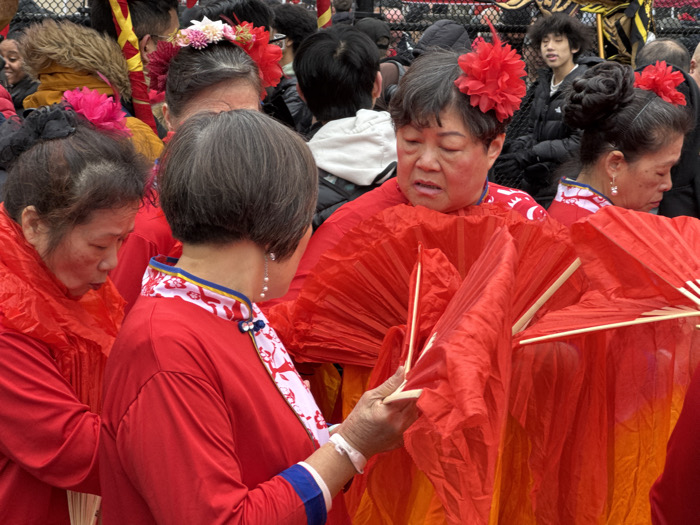 The image captures a vibrant scene, likely outdoors, featuring a group of predominantly older Asian women dressed in striking red attire, engaged in what appears to be a cultural performance or parade. The overall impression is one of festivity and coordinated activity.
In the foreground and mid-ground, five women are prominently featured. The woman in the center foreground has her back mostly to the viewer, revealing short, neatly cut gray hair and a pearl drop earring. She wears a bright red tunic with an ornate high collar featuring white fabric patterned with blue and red motifs, trimmed with a golden stripe and a blue band. She holds a closed red fan in her hands. To her left, another woman, partially visible, has dark hair with a pink flower decoration and is also clad in red. She appears to be looking down or adjusting something.
The central figure in the image is an older woman facing right, her expression appearing somewhat pensive or possibly speaking. She has short, dark, graying hair adorned with a large, ruffled red flower. Her red top has a patterned border visible near her neck. She is actively holding a large, partially open, pleated red fan made of fabric, with light-colored wooden or bamboo spokes. Another similar fan is visible behind hers.
To the far right, another woman is seen in profile, looking down. She has dark hair styled in two elaborate buns, each decorated with a vibrant red flower, and wears pearl drop earrings similar to the woman with gray hair. She is also dressed in a red garment. Behind her, another woman's face is partially visible.
In the background, other people can be seen, suggesting a crowd. A young man with dark, curly hair wearing a black puffer jacket is visible looking towards the right. A dark metal fence is behind the participants, indicating an outdoor, public setting. The lighting suggests it is daytime, possibly overcast, creating soft shadows. The dominant color throughout the image is a rich red, associated with good fortune and celebration in many Asian cultures. The women appear to be either preparing for, waiting to perform, or actively participating in a coordinated display involving the fans.