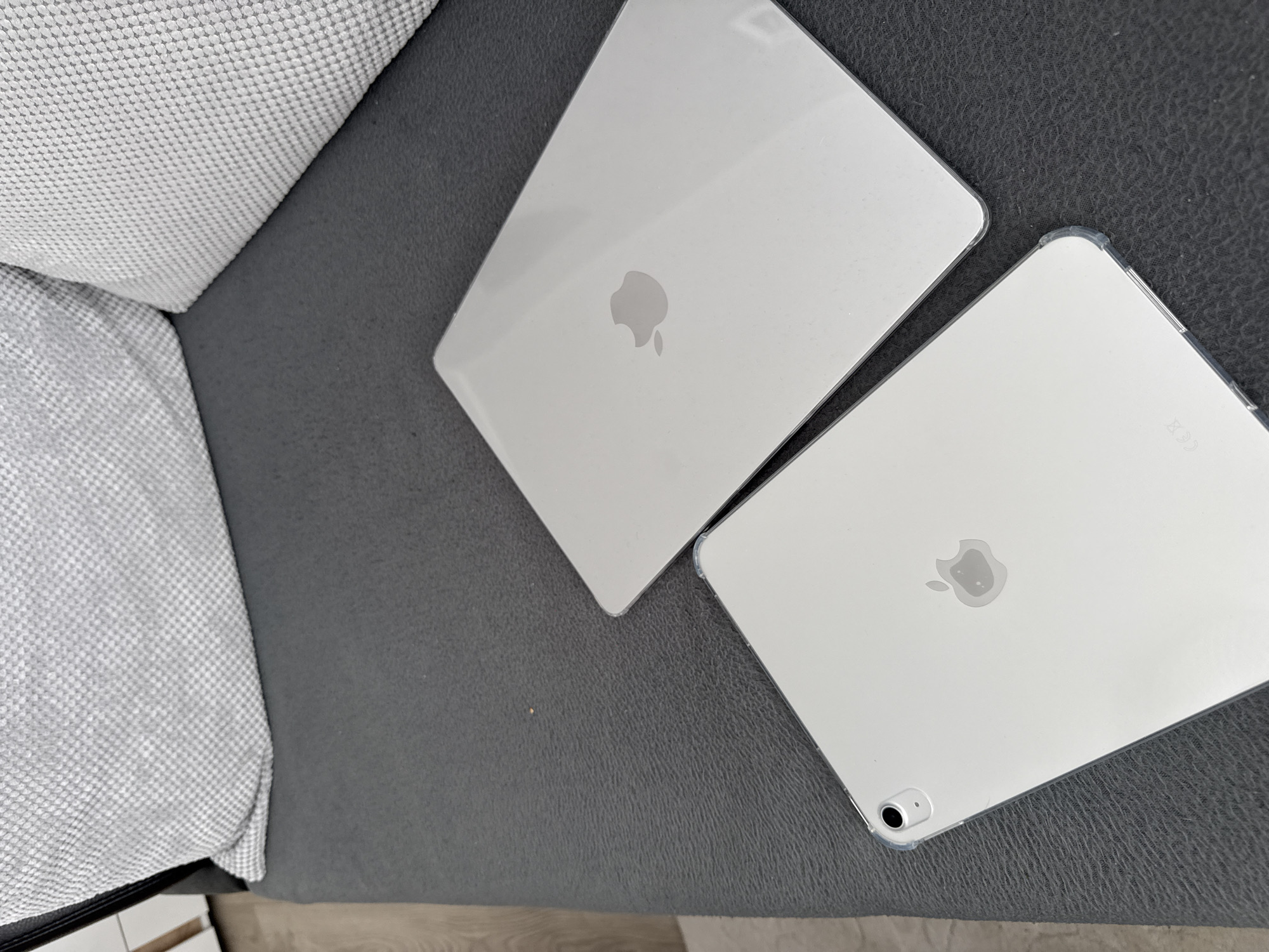 The image shows two silver Apple iPads lying on a gray surface, possibly a couch. One iPad has a clear protective case on the corners. The backdrop includes a gray patterned cushion or piece of furniture. The scene does not appear to be in the city of Lovech, Bulgaria, as there are no identifiable elements linking it to that location.
