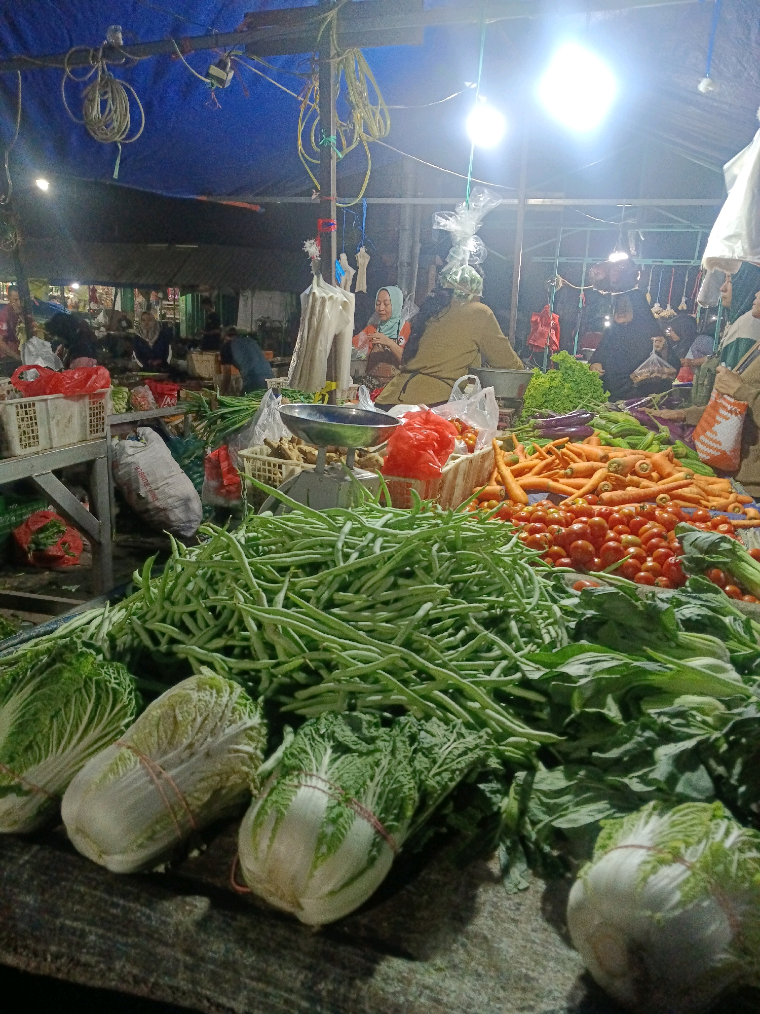 This is a medium shot, taken from a slightly low angle, of a produce stand at an outdoor market in Wonocolo, Indonesia. The market appears to be covered by a blue tarp, with bright lights illuminating the scene. In the foreground, a large assortment of fresh vegetables is displayed on a dark surface. Prominently featured are bunches of green beans, several heads of napa cabbage tied with red bands, and leafy greens. Behind these, a variety of other vegetables are visible, including carrots, tomatoes, and what appear to be eggplants. In the background, several people, some wearing headscarves, are tending to the stall and interacting with customers. Bags of produce and market baskets are scattered throughout the scene. The overall impression is that of a busy, vibrant market stall.
