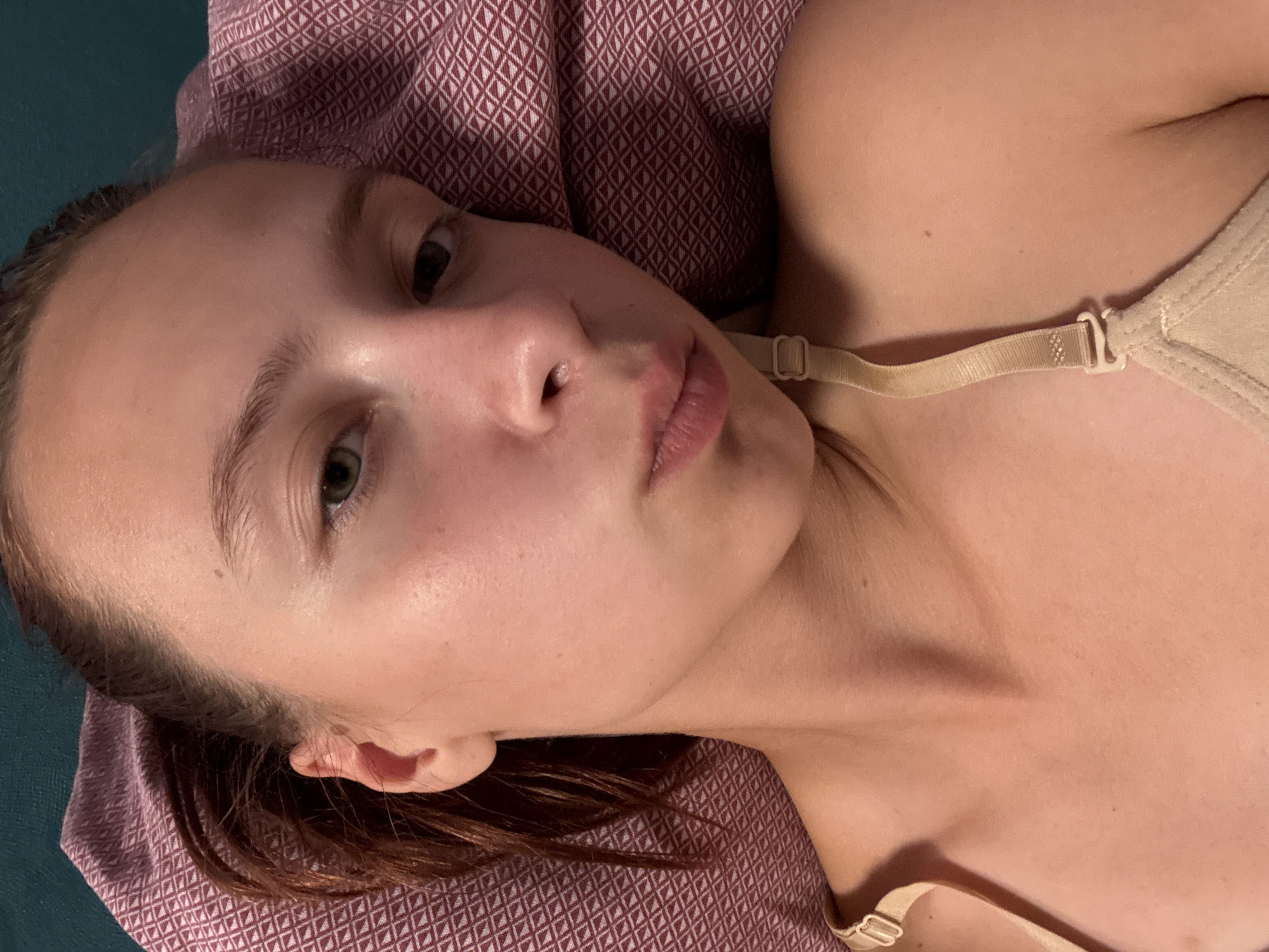 A young woman with her hair pulled back in a ponytail is lying on a pillow with a patterned surface. She is wearing a beige bra with a strap visible on her shoulder. The woman is looking towards the camera with a neutral expression. The background is a teal color.