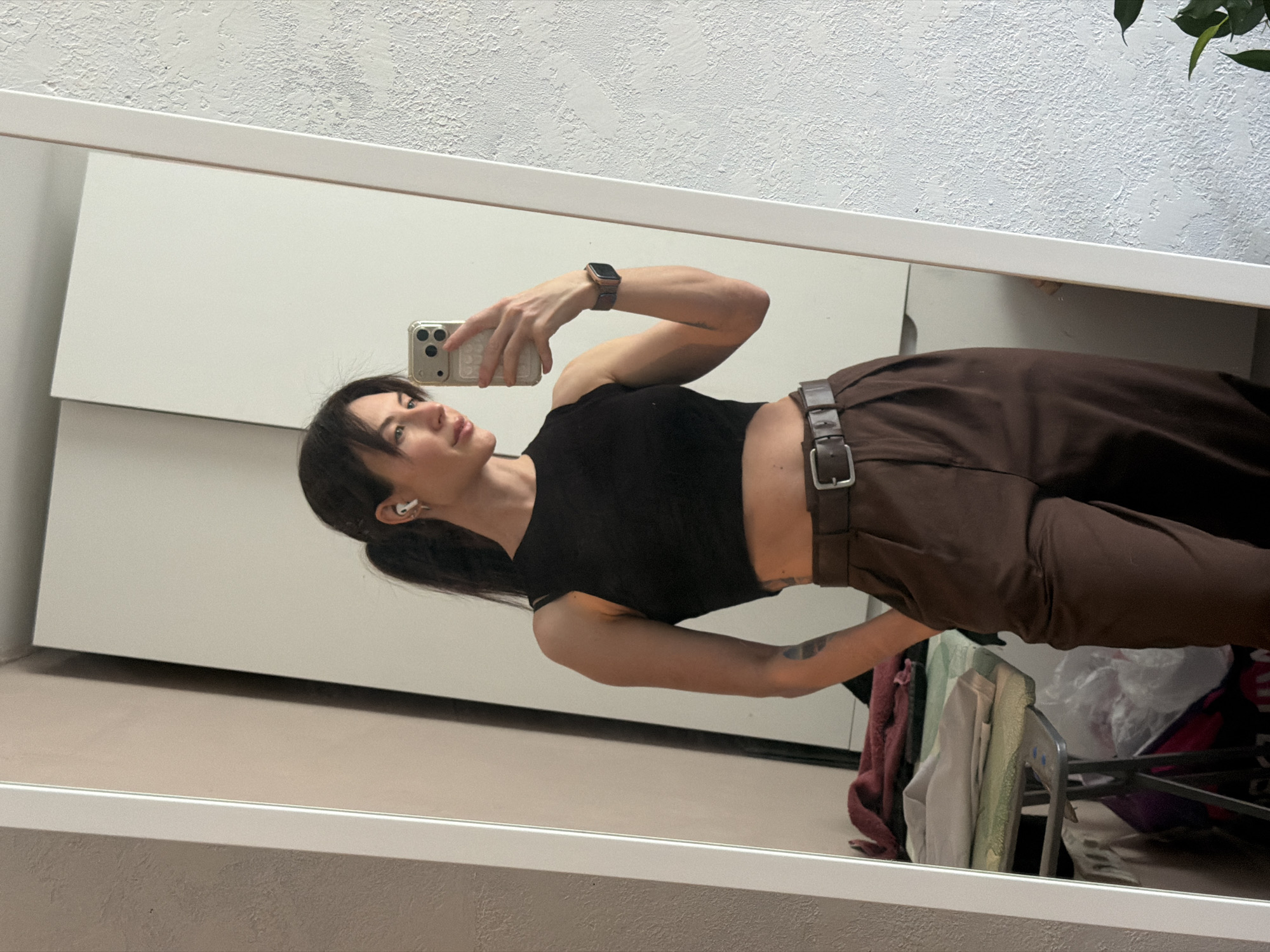 A woman is reflected in a mirror, taking a selfie with a smartphone. She is wearing a black cropped top and brown pants with a belt. She has dark hair pulled back in a ponytail and is wearing a watch and an earbud. The reflection shows a white wall and a shelf with items that suggest a cluttered or storage area. The reflection suggests an indoor setting in what seems to be a room. The location, Perm, Russia, is not evident within the provided visual information.
