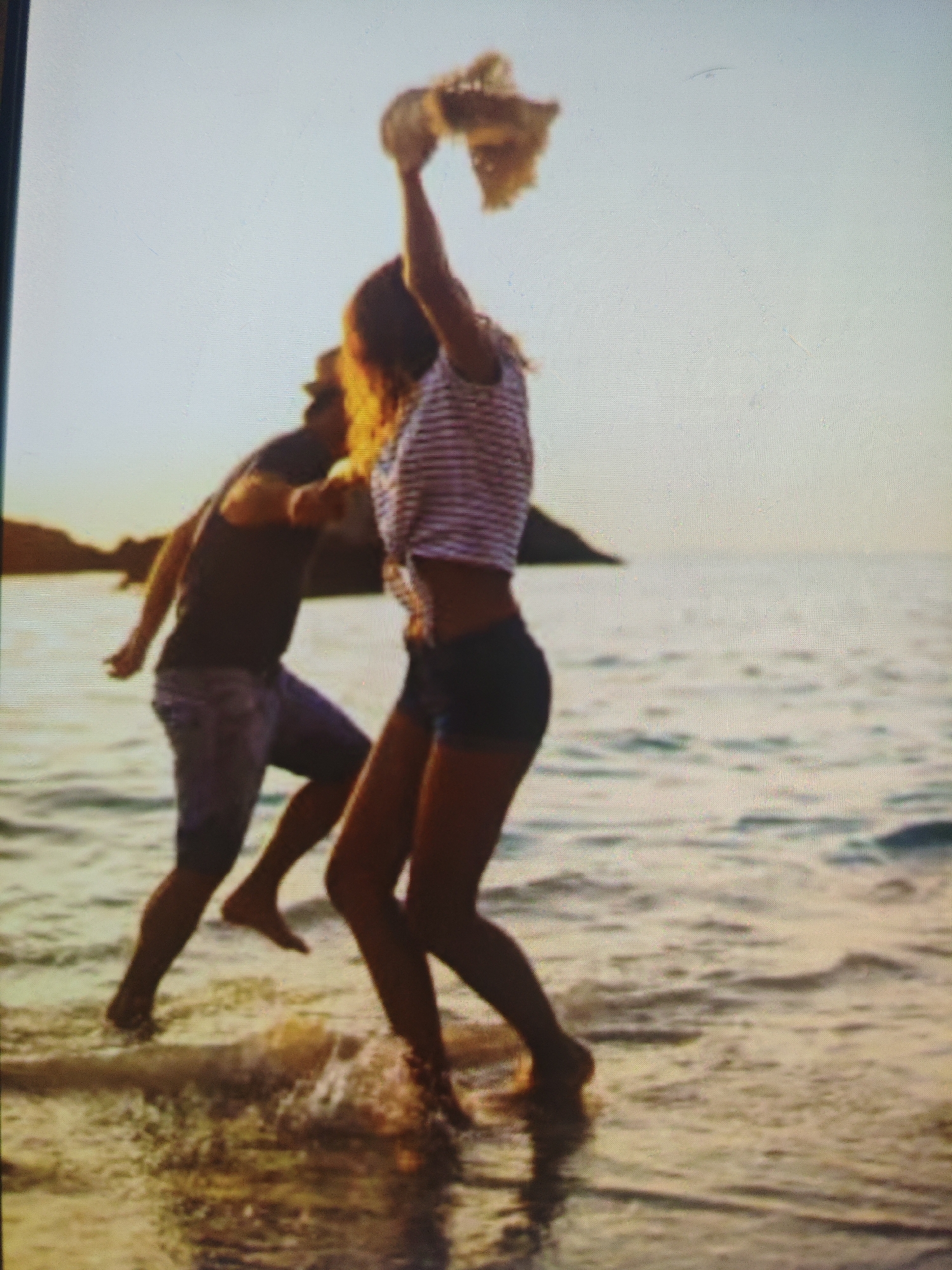 The image depicts two individuals, a male and a female, interacting on a beach. The female is positioned in the foreground, facing away from the camera and towards the ocean. She is wearing a striped crop top and dark shorts, with her right arm raised, holding a straw hat. The male is behind her, facing her, wearing a dark t-shirt and denim shorts. Both are wading in shallow water. The background shows a body of water, likely the sea, with a faint outline of land in the distance. The lighting suggests it is either dawn or dusk, with a soft, warm glow. The image is pixelated and somewhat blurry, making precise details difficult to ascertain. The scene is not located in Krasnaya Polyana, Russia, as this is a mountain resort town and not a coastal location.