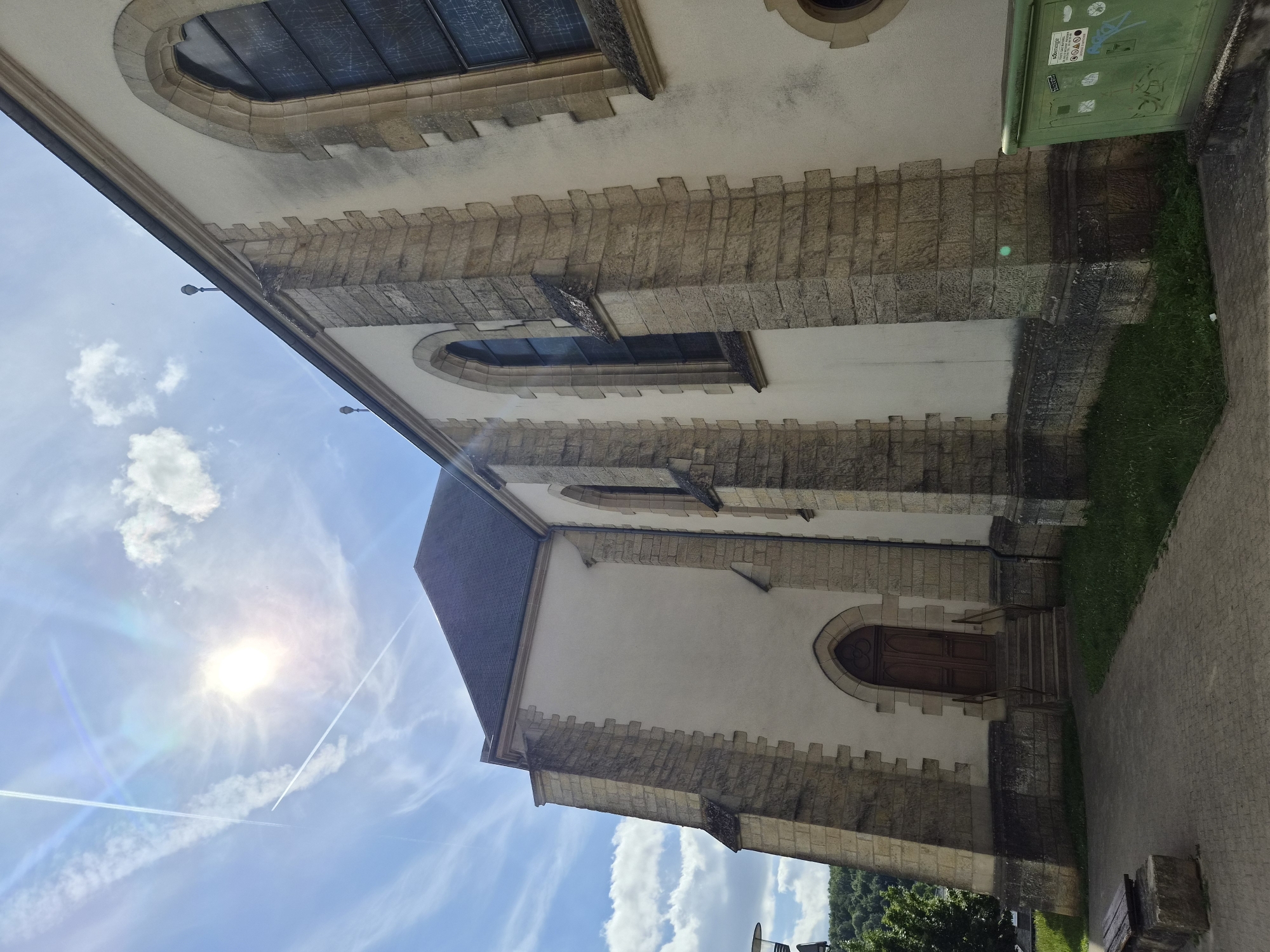 The image depicts the exterior of a stone and stucco church, likely Saint-Alban's Church, in Differdange, Luxembourg. The building features arched windows and a dark tiled roof. Sunlight streams from the upper left, creating lens flare and casting shadows. Two contrails are visible in the bright blue sky with scattered white clouds. In the foreground, a paved walkway leads towards the church entrance, which has a wooden door and a set of stairs. A dark wooden bench with stone supports sits to the left of the walkway. A green dumpster with stickers is positioned to the right of the church. The environment appears to be a public space with some greenery and a hillside in the background.